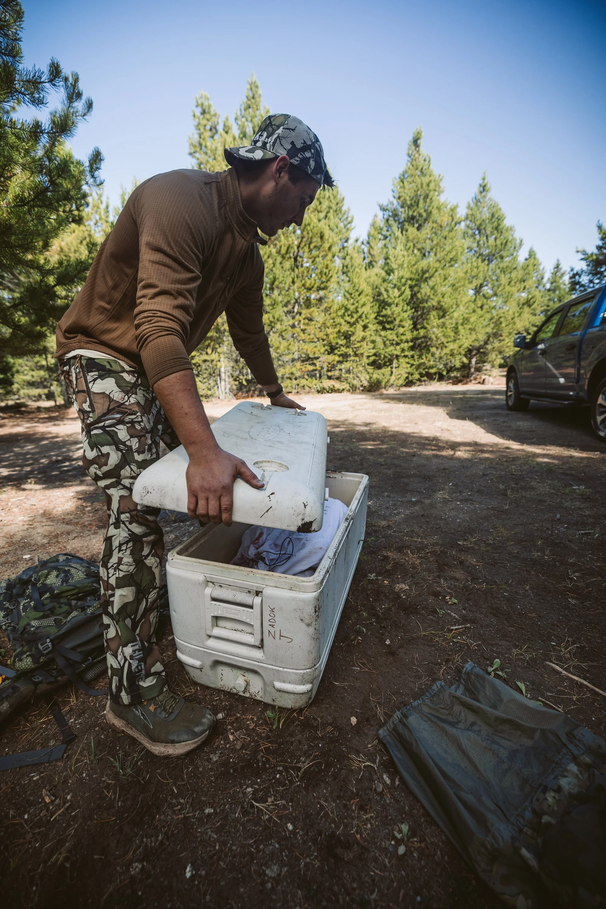 Man in camouflage clothing opening a large cooler outdoors in a forested area next to a vehicle.