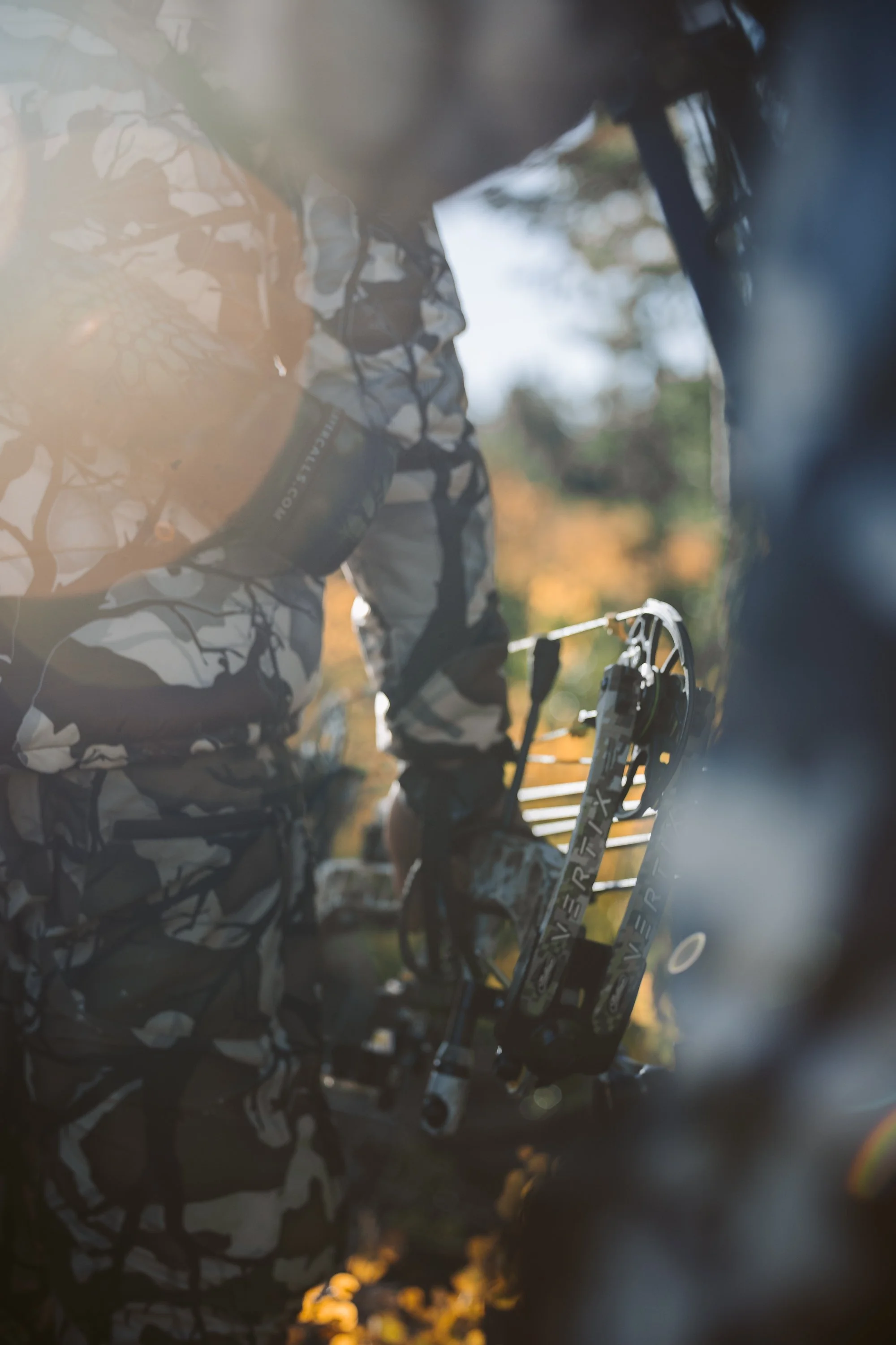 Person in camouflage holding a hunting bow outdoors, with sunlight in the background.
