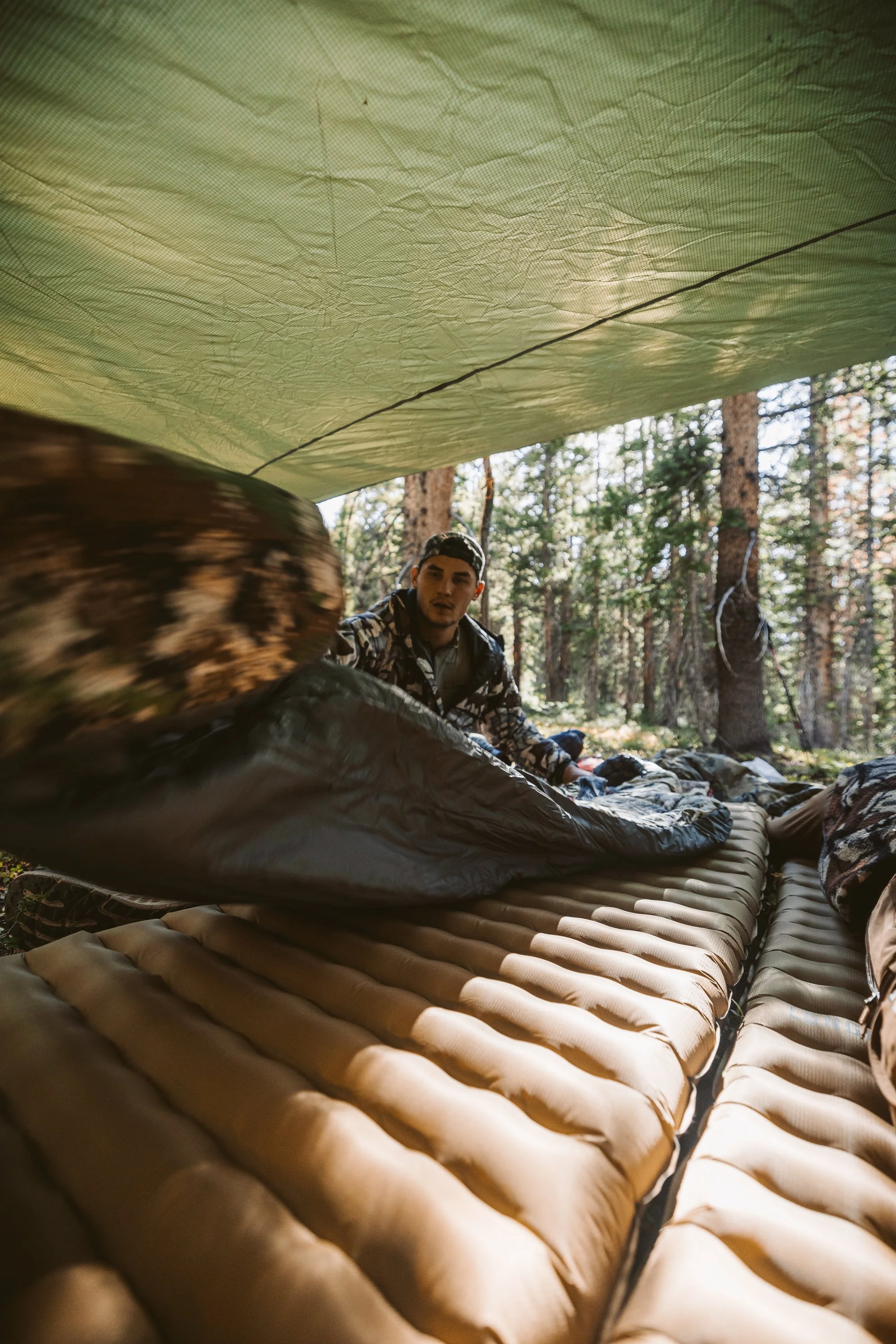 Person setting up a sleeping area under a tarp in a forest, using a sleeping pad and sleeping bag.
