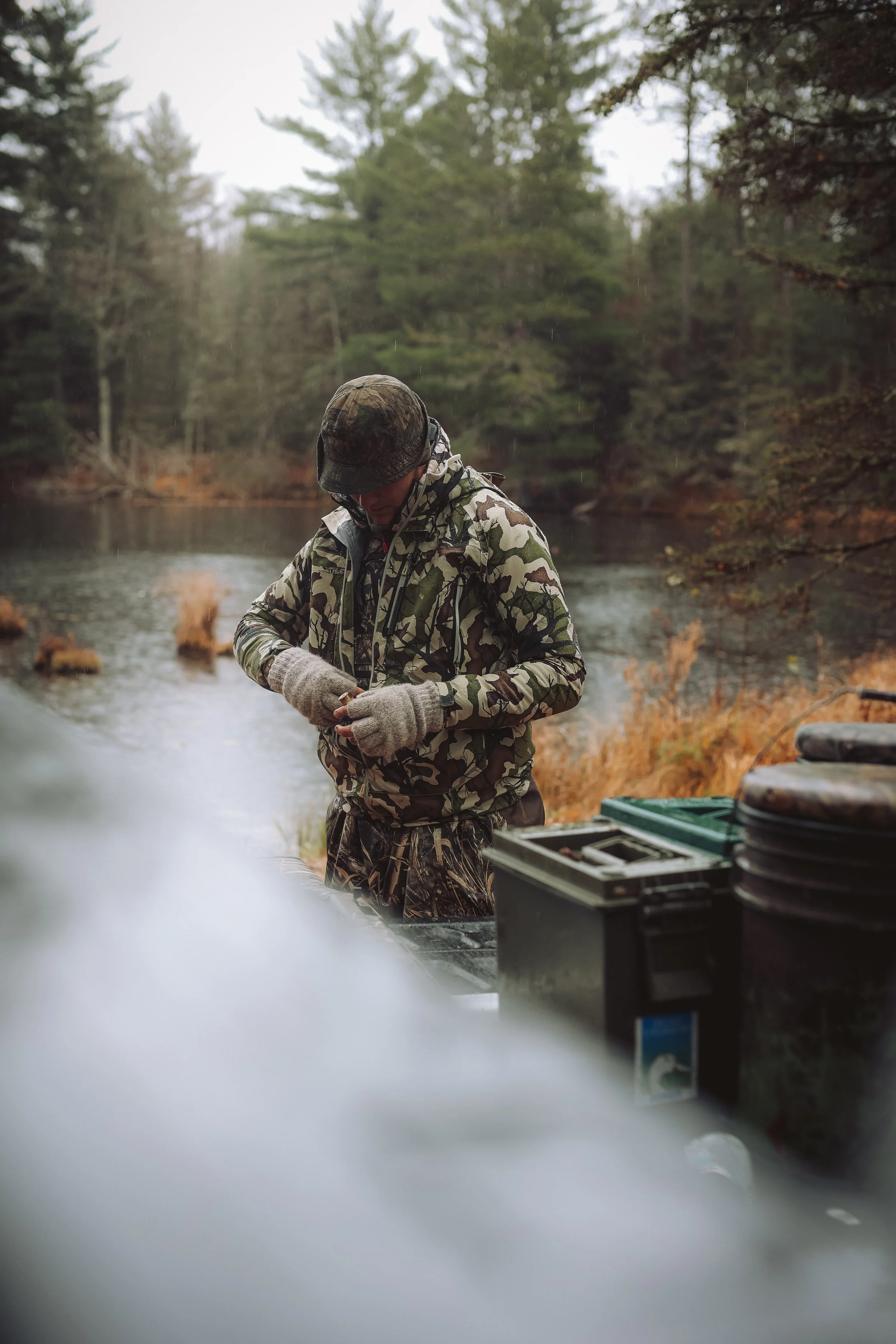 Person in camouflage gear standing near a lake with evergreen trees and outdoor equipment.