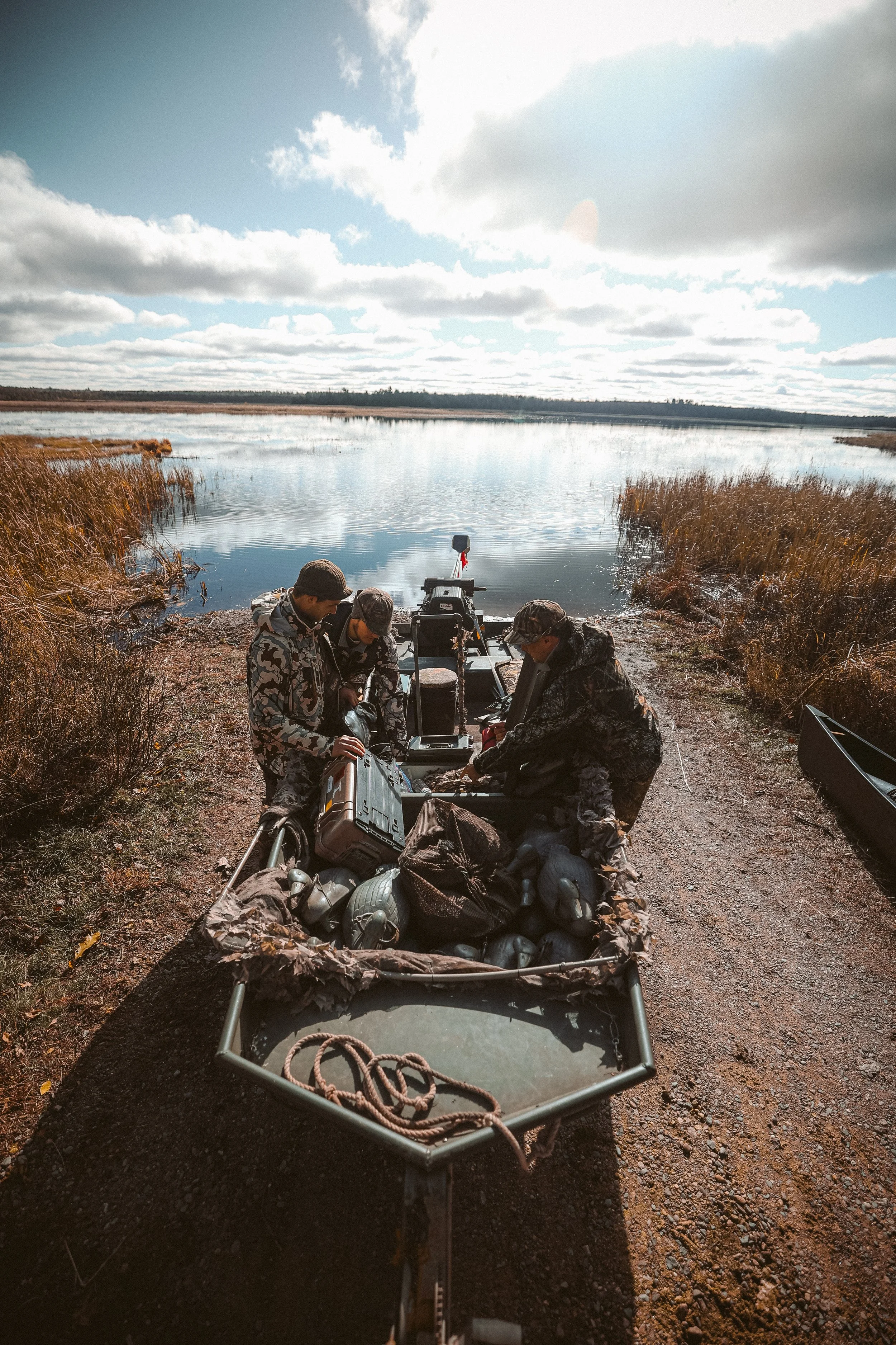 Three people in camouflage gear prepare a boat with hunting equipment, including decoys, near a scenic marshland under a partly cloudy sky.