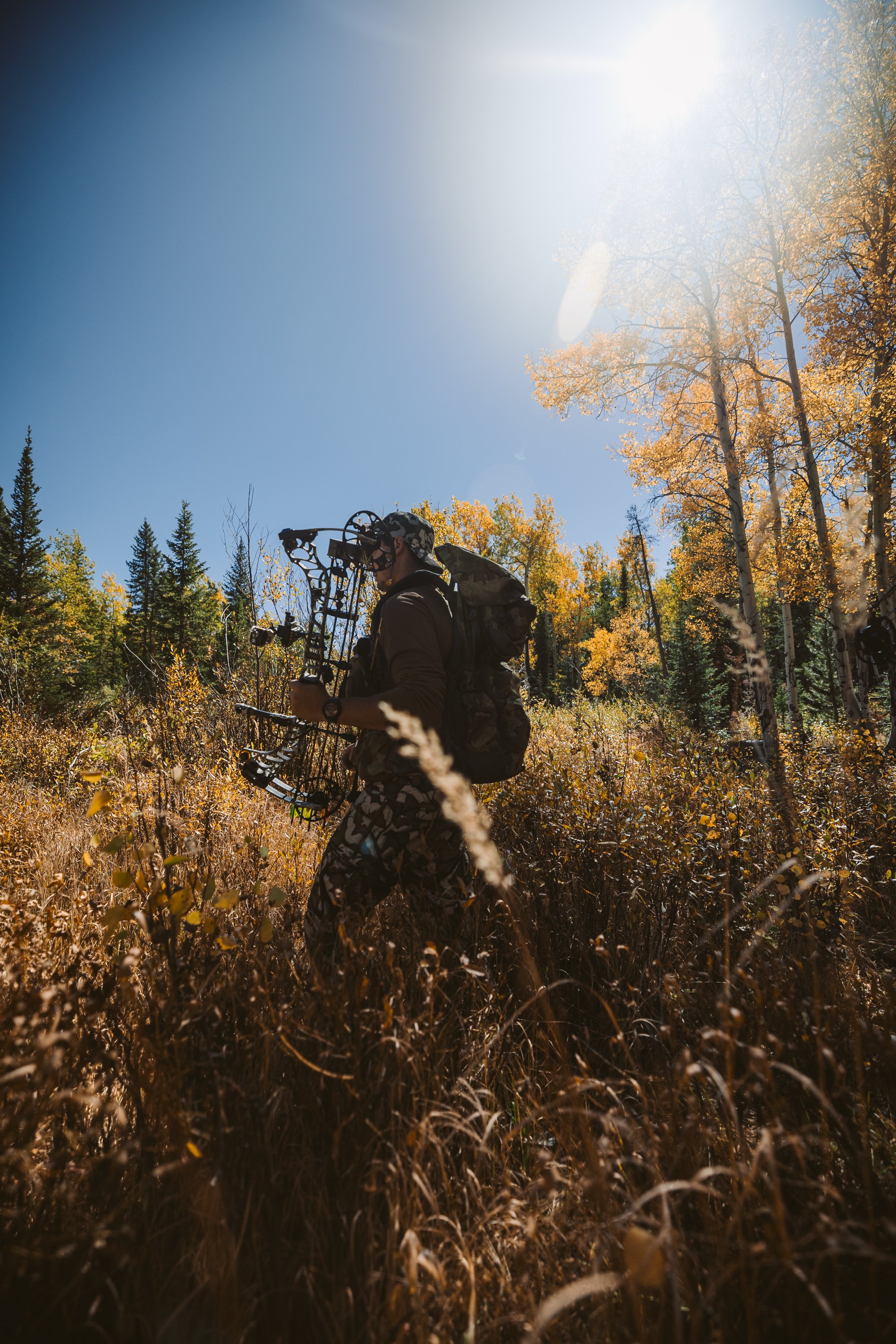 Archer walking through autumn forest, holding a compound bow with backpack, under bright sunlight.