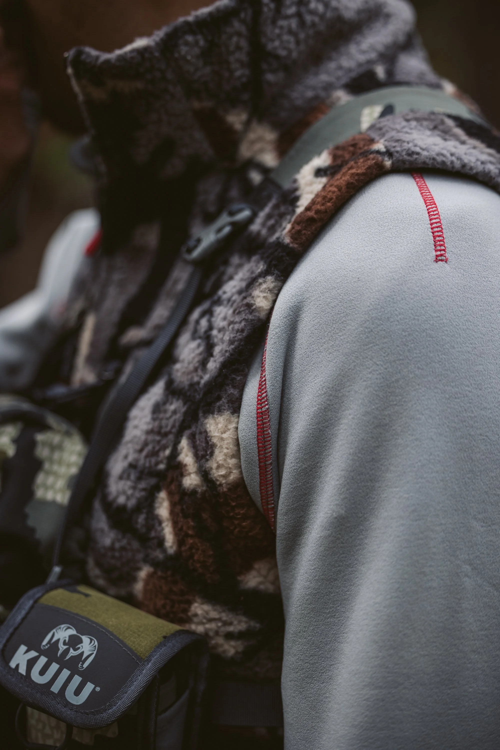 Close-up of outdoor clothing featuring a gray fleece jacket, patterned vest, and KUIU branded gear pouch.