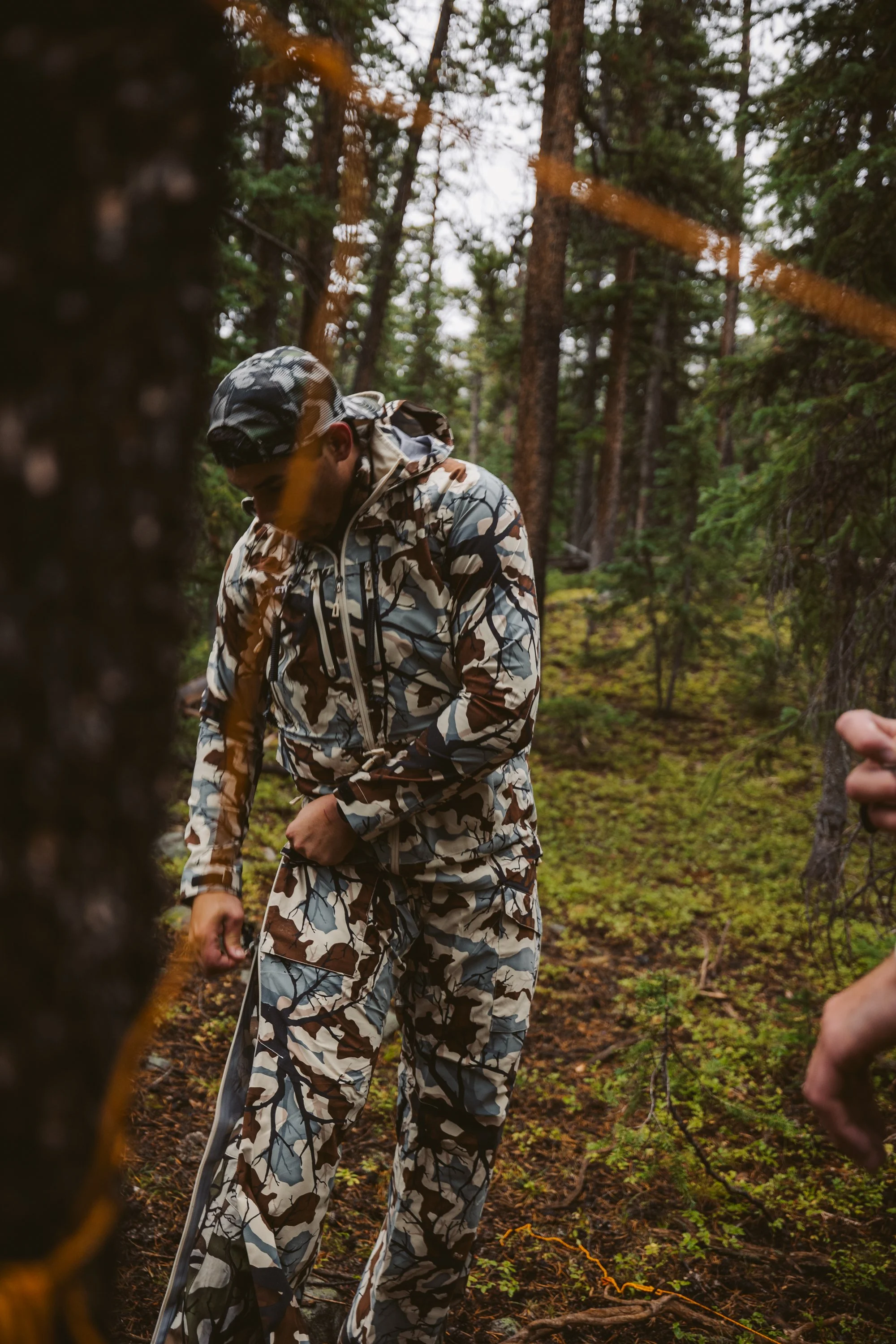Person in camouflage clothing outdoors in a wooded area, adjusting clothing near trees.