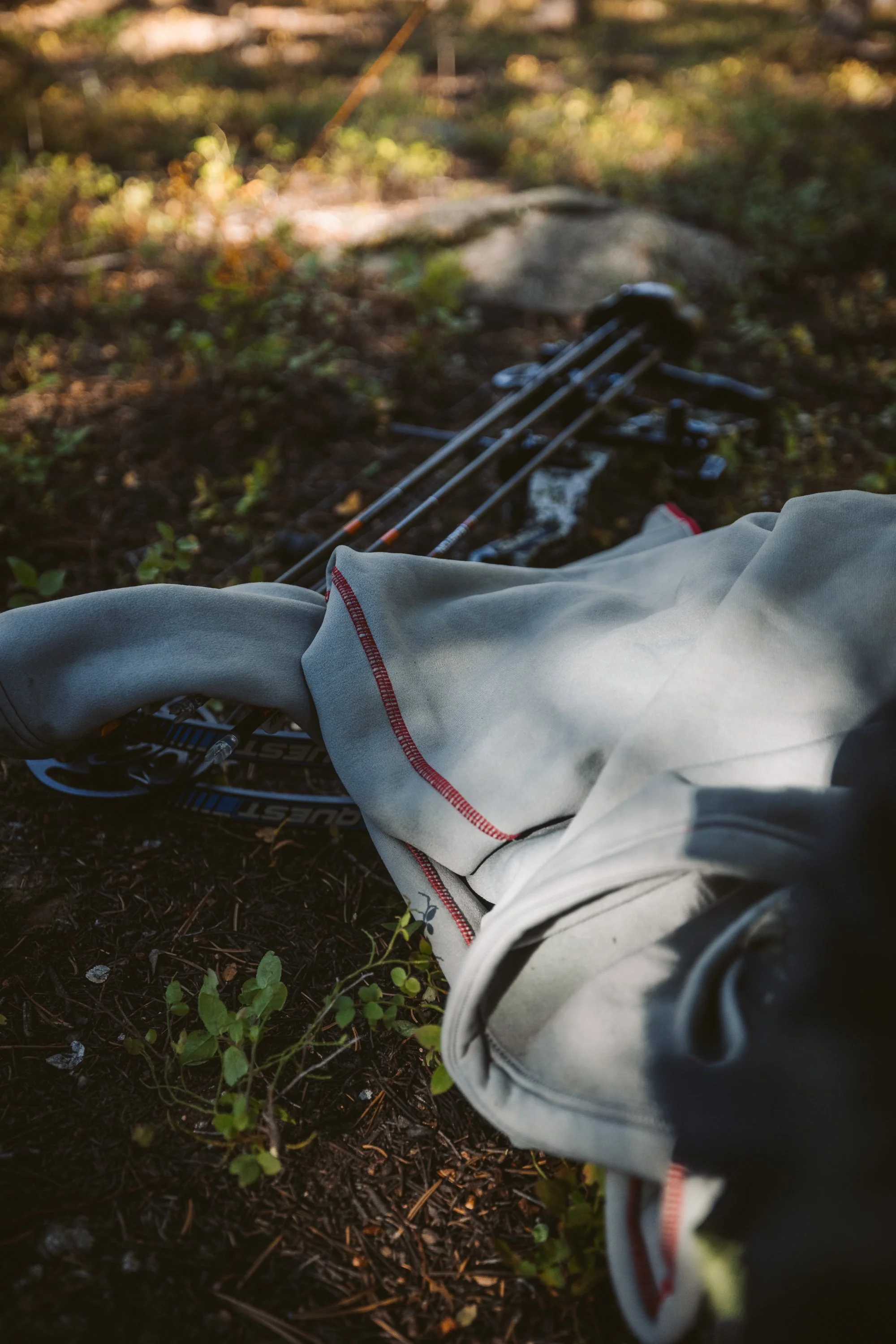 A gray sweatshirt with red stitching is laid over a compound bow on the forest floor, surrounded by greenery and sunlight.
