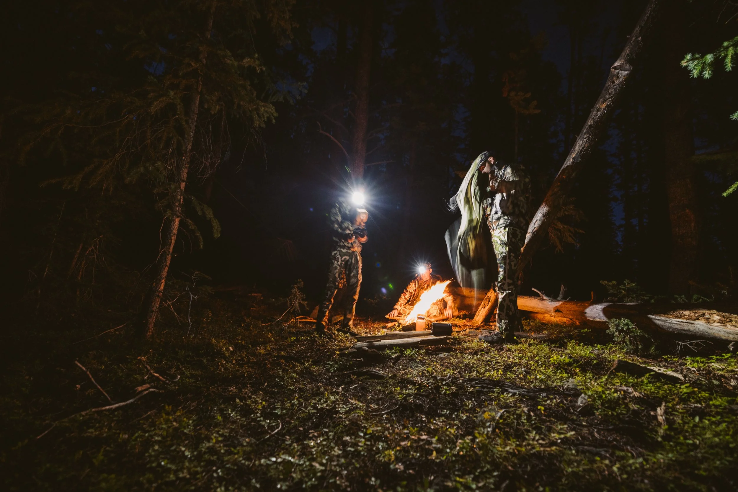 Two people camping in a forest at night, wearing headlamps, with a campfire illuminating the scene.