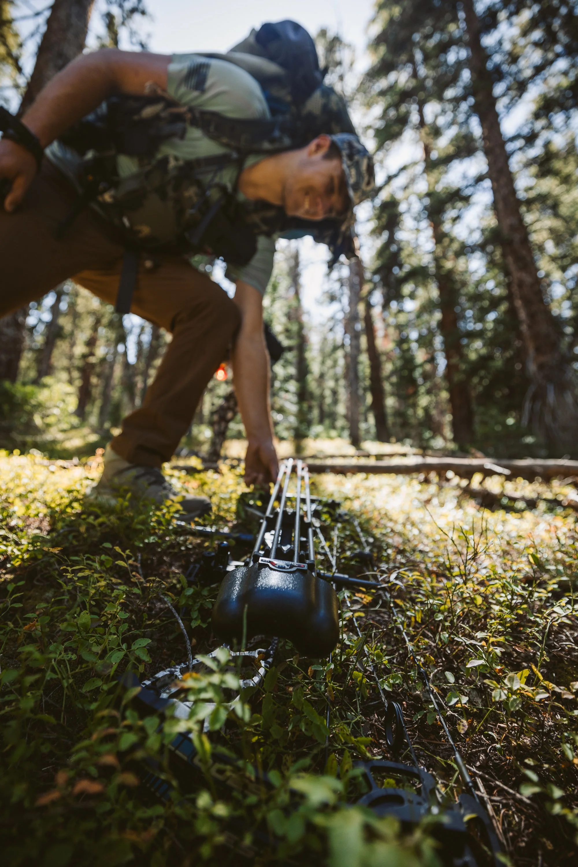 A person with a backpack and camo clothing picking up a compound bow in a forest.