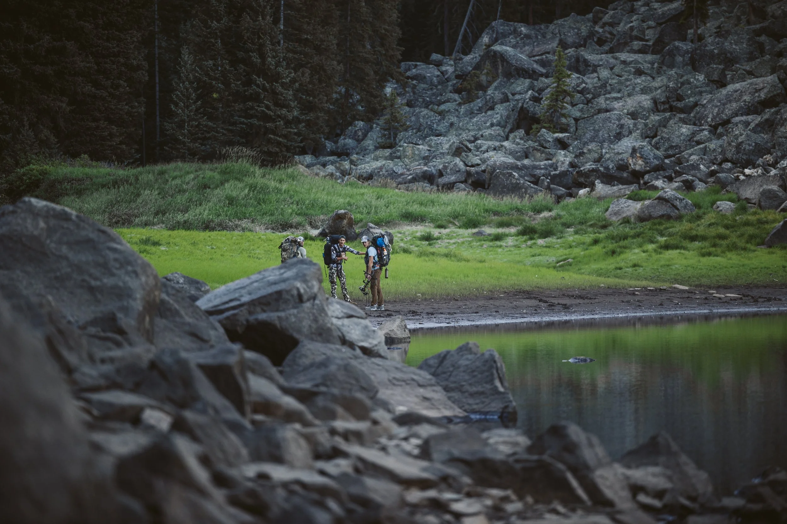 Three hikers with backpacks standing near a forested lakeshore surrounded by rocks and greenery.