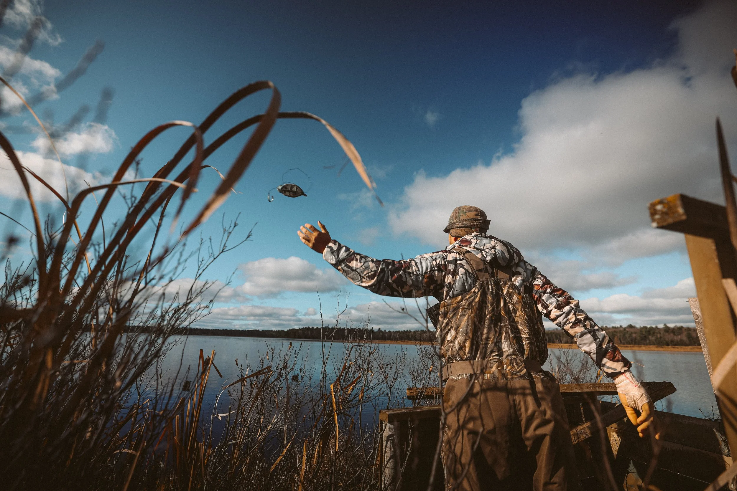 Person in camouflage clothing throwing a duck decoy by a lake, surrounded by reeds, under a cloudy blue sky.
