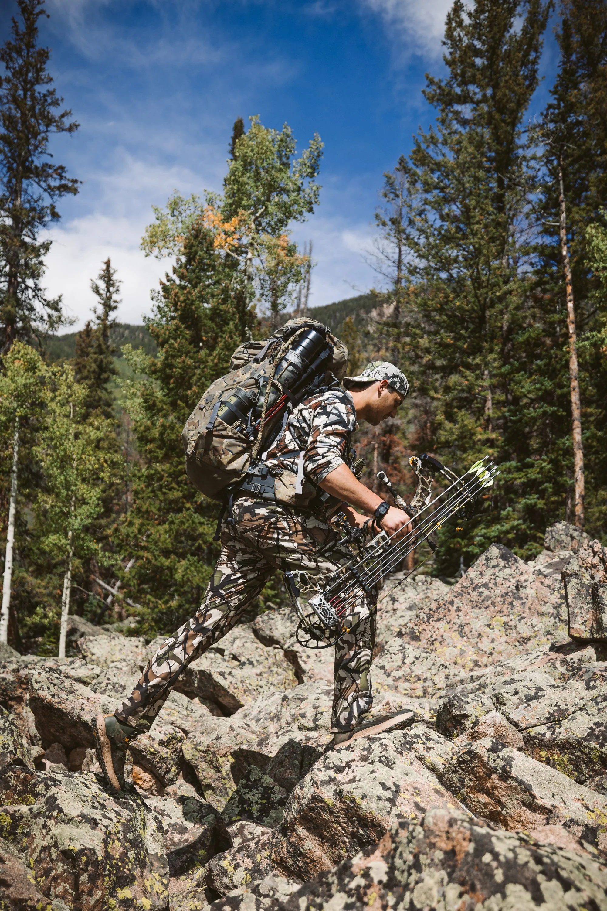 A person in camouflage gear hiking over rocky terrain, carrying a bow and arrows, with a backpack. The background features a forest with tall trees and a blue sky.