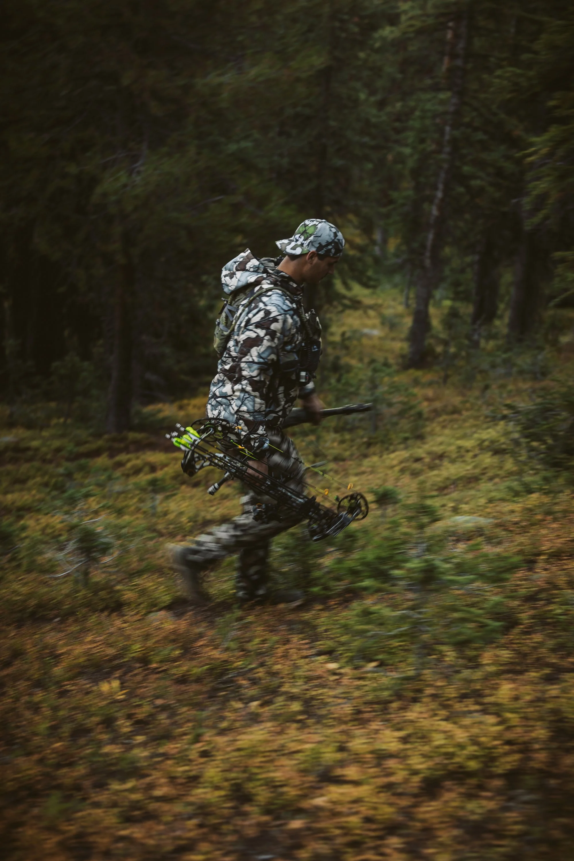 A person wearing camouflage gear and holding a compound bow walks through a forest.