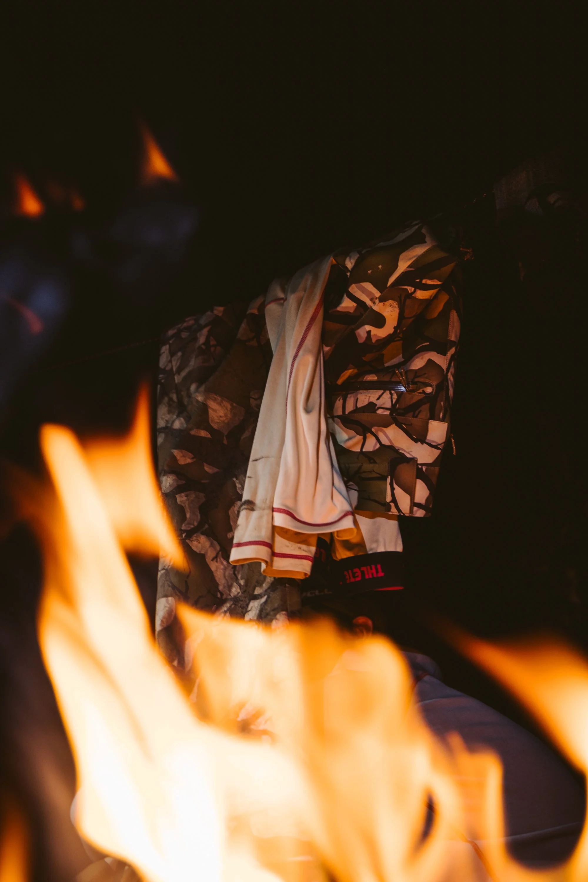 Clothes hanging near a campfire with visible flames, including a camouflage jacket and white fabric.