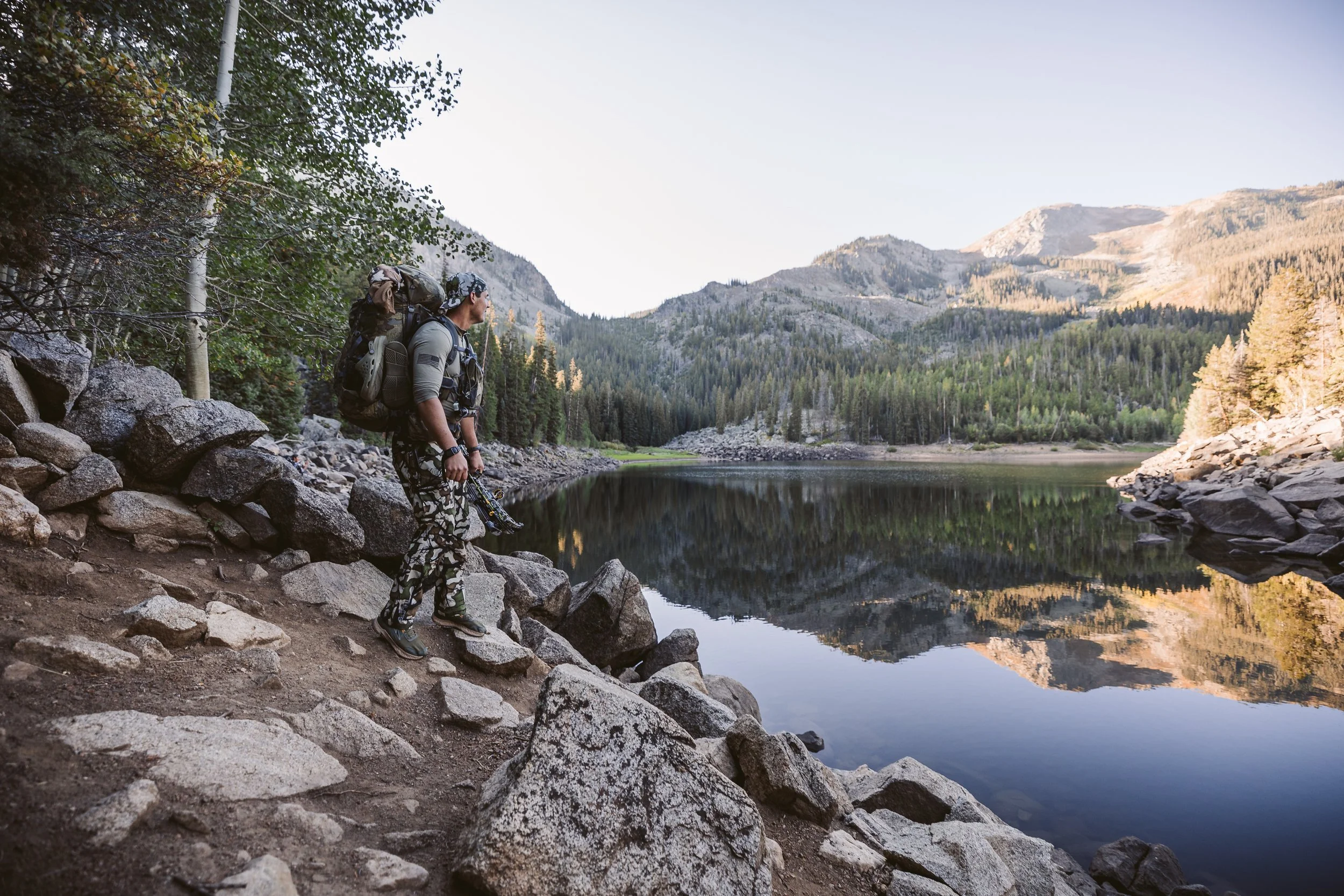 Person hiking near a mountain lake with rocky terrain; trees and mountains are reflected in the water.
