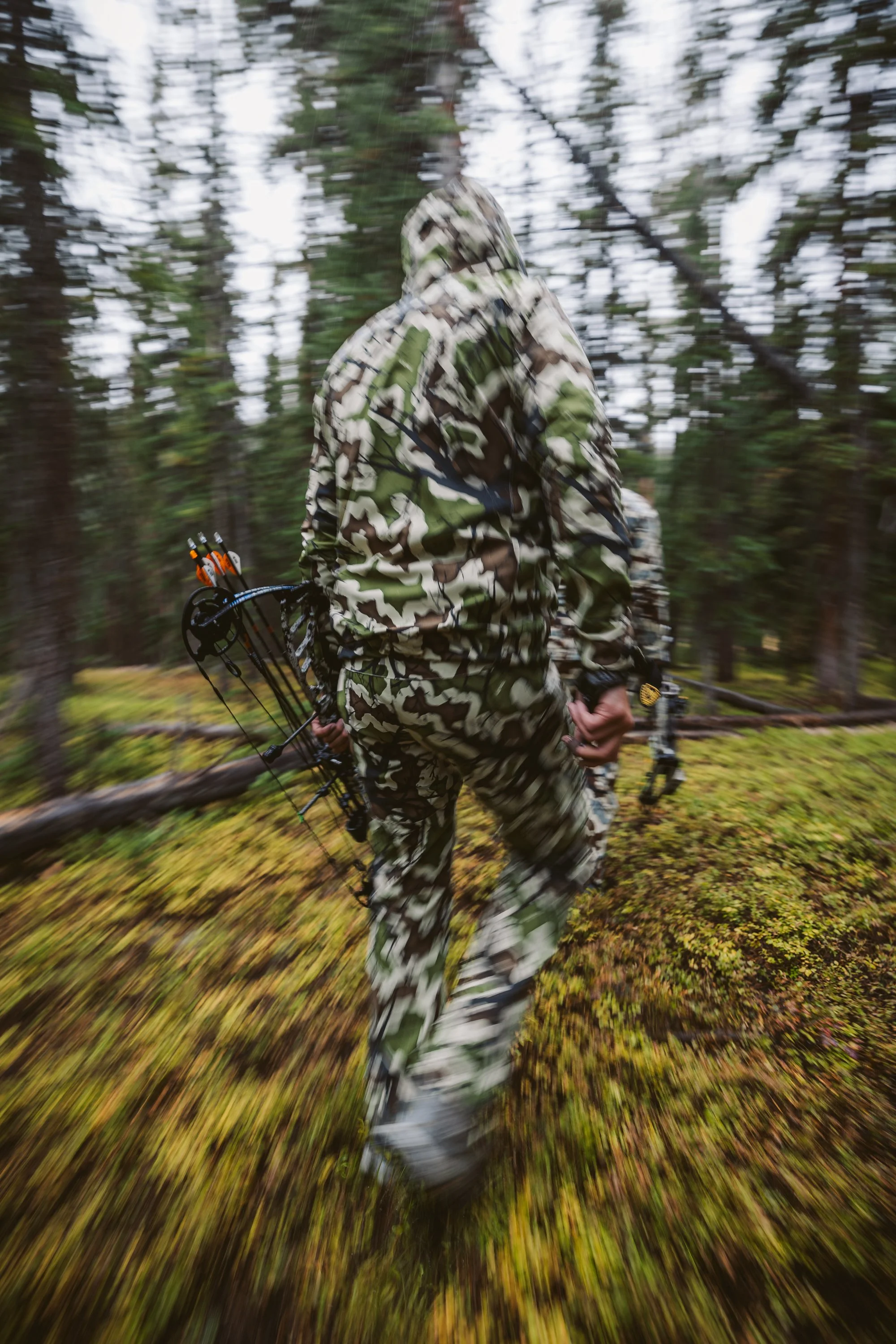 Person in camouflage with a bow walking through a forest.