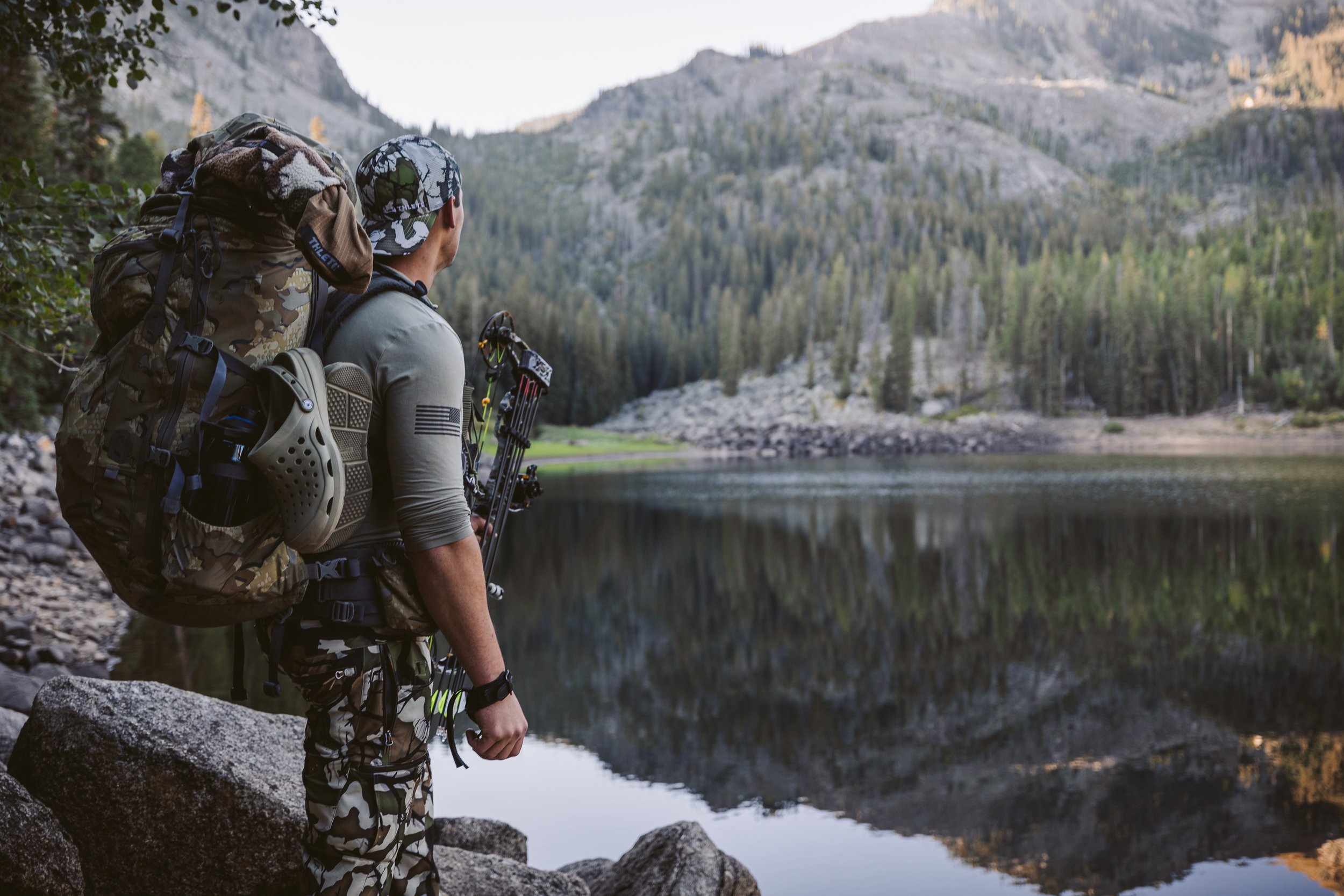 Person in camo gear with a backpack and bow standing by a lake with forest and mountains in the background.
