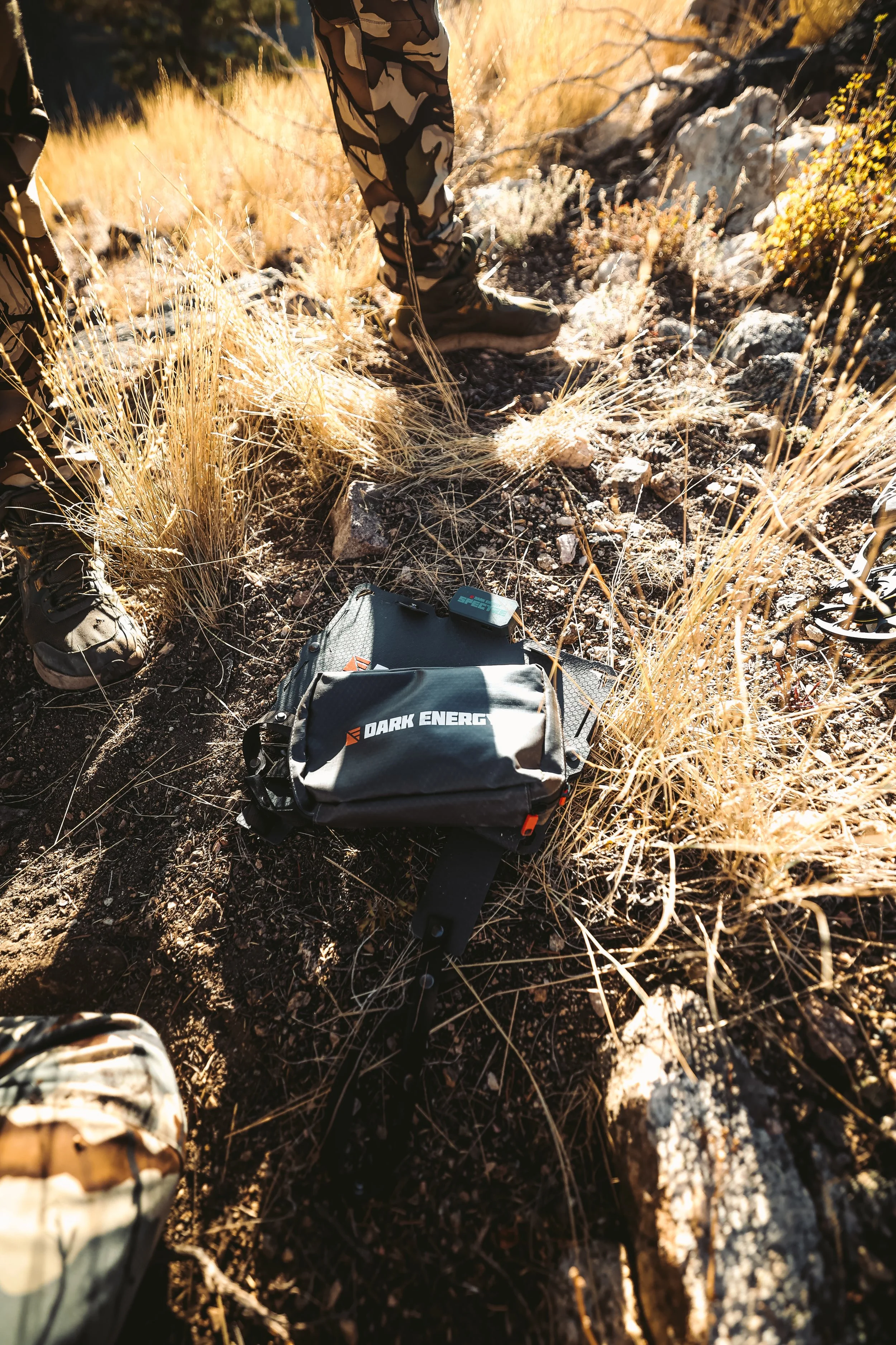 Person wearing camouflage pants and boots standing next to a bag labeled 'Dark Energy' on rocky terrain with dry grass.