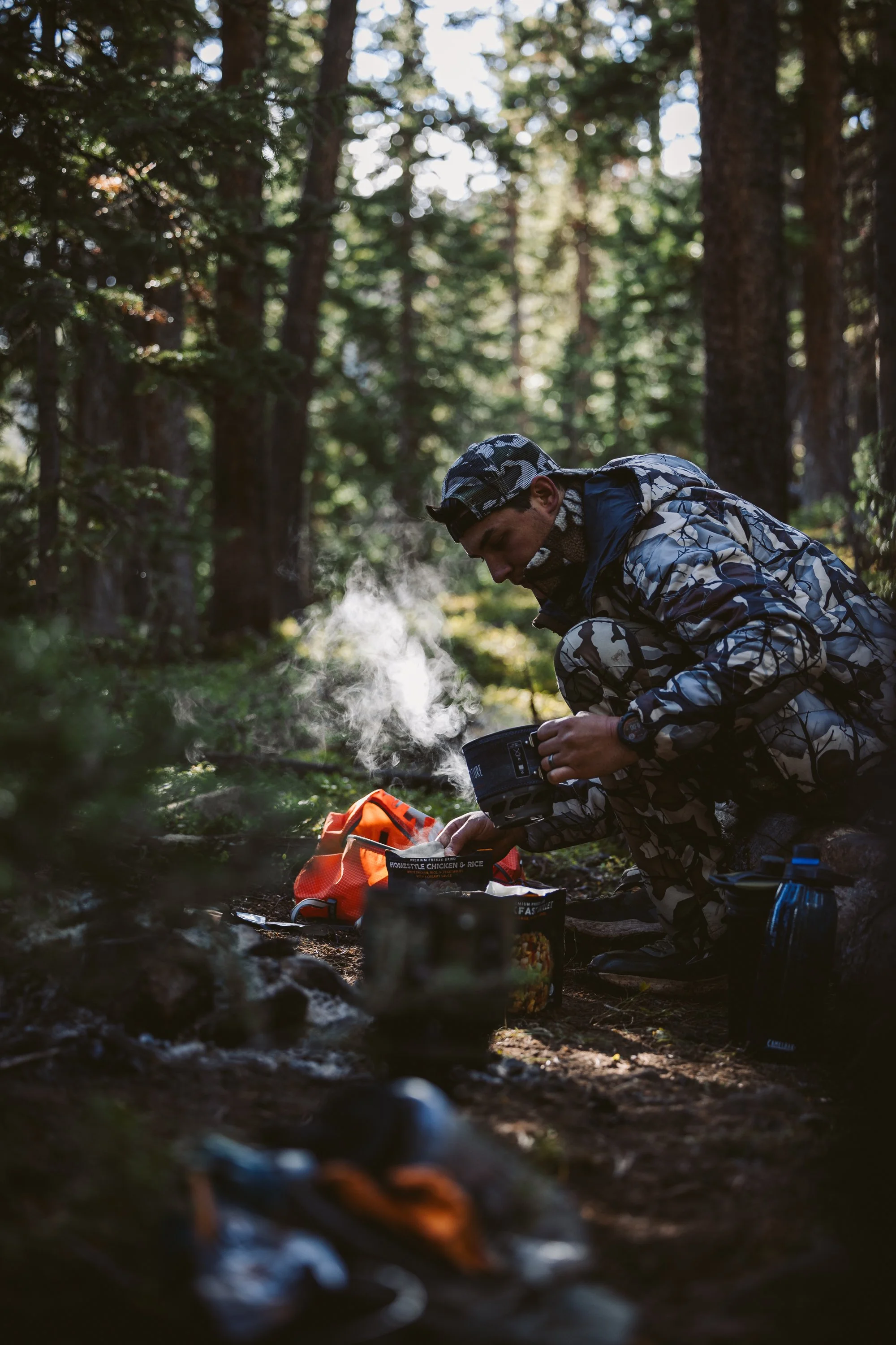 Person preparing a meal in a forest setting, wearing camouflage clothing. Steam is rising from a cooking pot on the ground surrounded by camping gear and food packets.