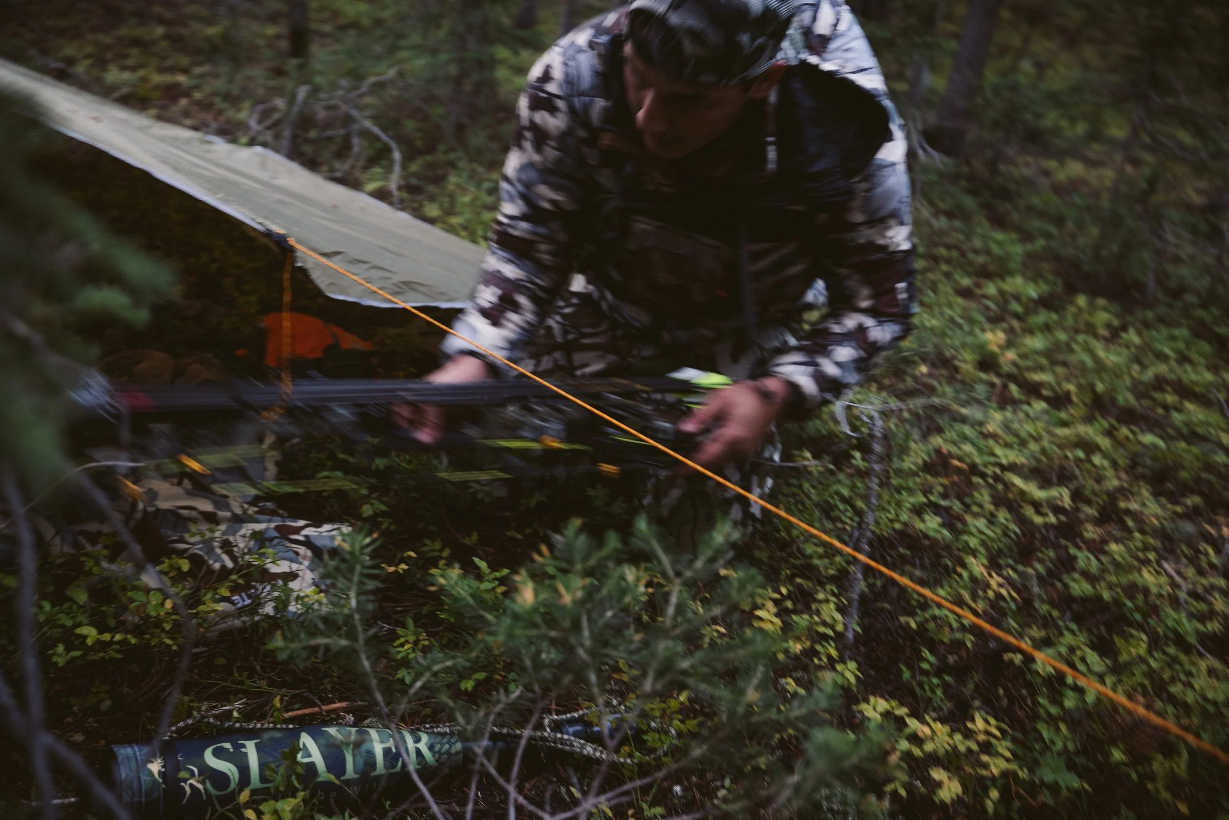 Person in camouflage holding bow near a tent in a forested area.