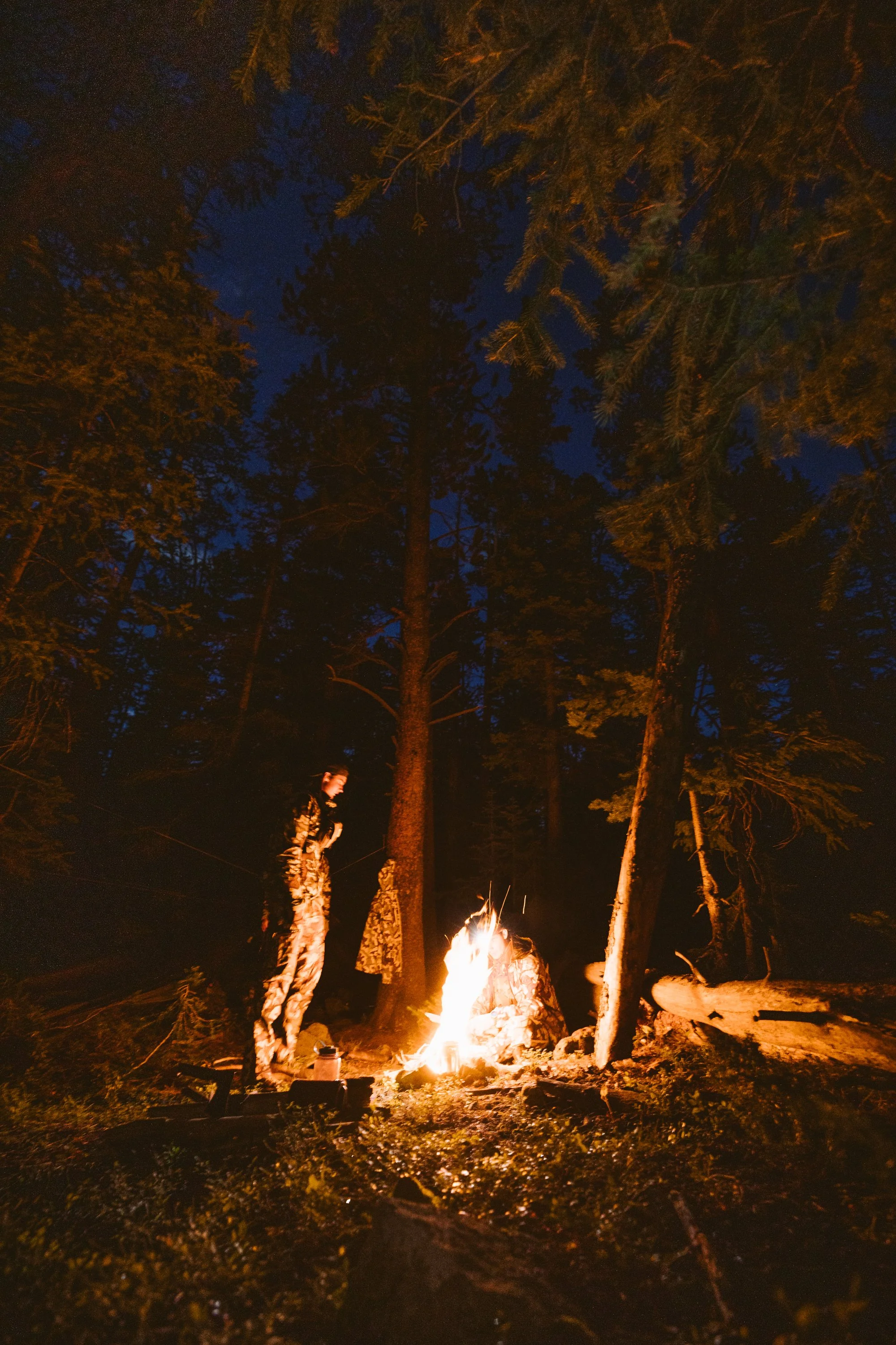 Two people in camouflage by a campfire in a forest at night.