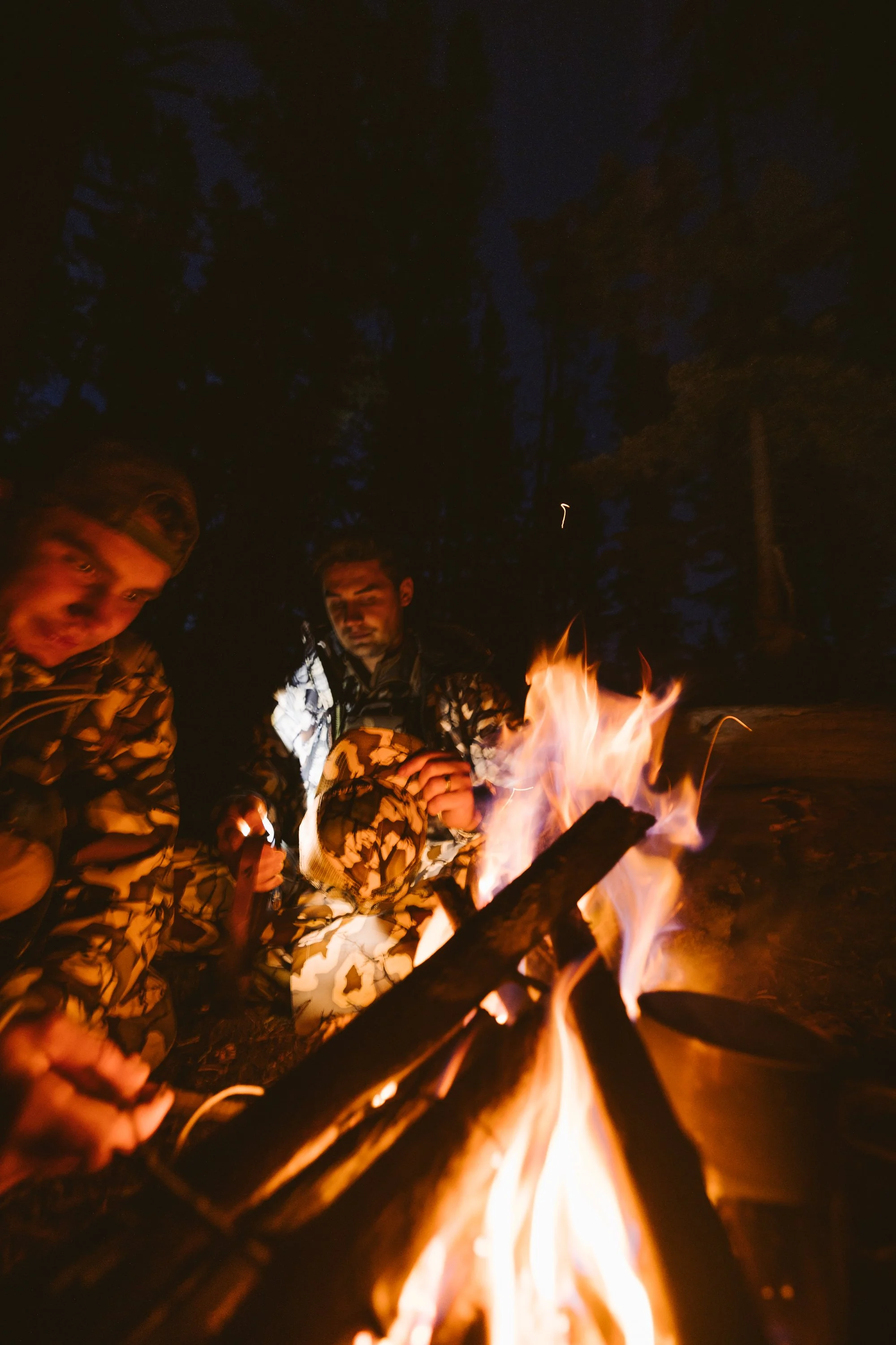 Two people in camouflage gear sitting by a campfire at night in a forest.