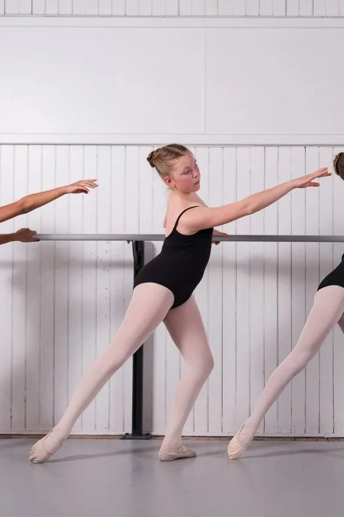 Young girl performing a pas de chat, wearing a purple and black leotard, inside a dance studio with pink and white walls and large windows.