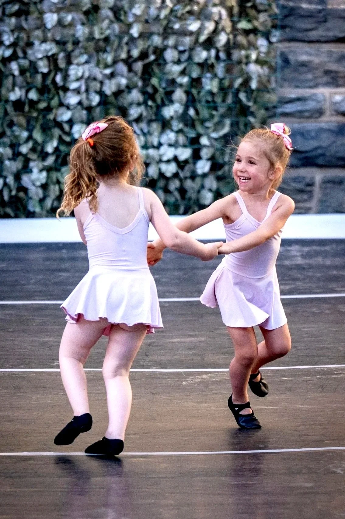 Young girl in a light pink ballet costume kneeling on the floor, smiling, with other young girls in ballet costumes sitting behind her on a stage or dance floor.