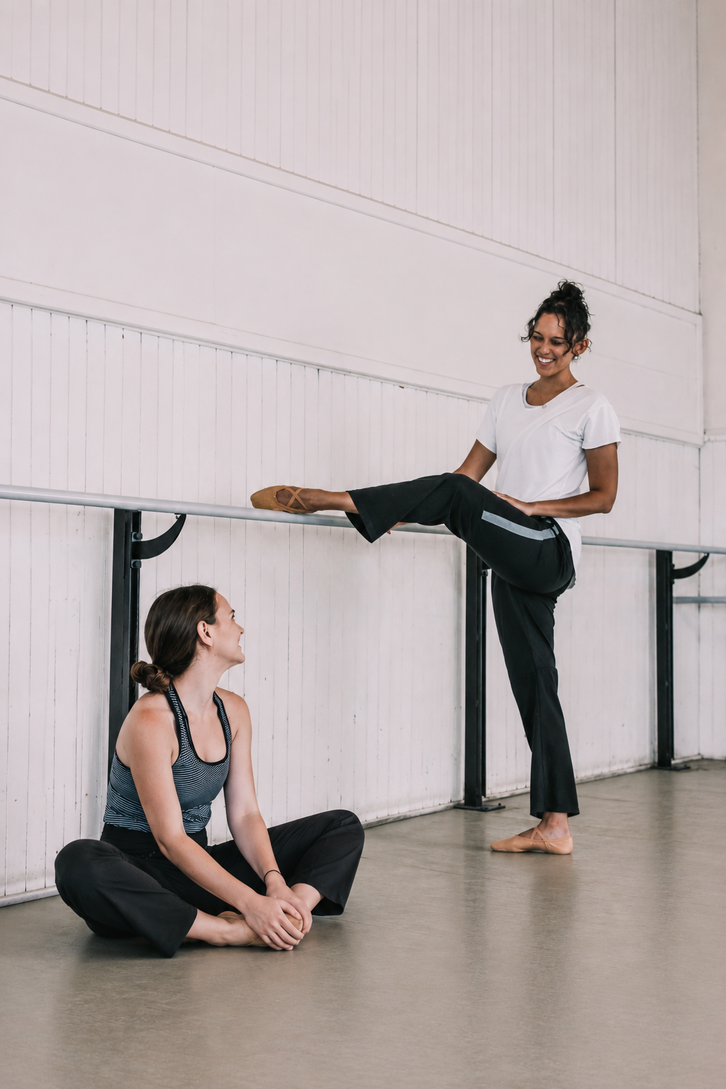 A ballerina practicing at a barre in a dance studio, smiling, wearing a white shirt and black pants, with one leg extended on the barre.