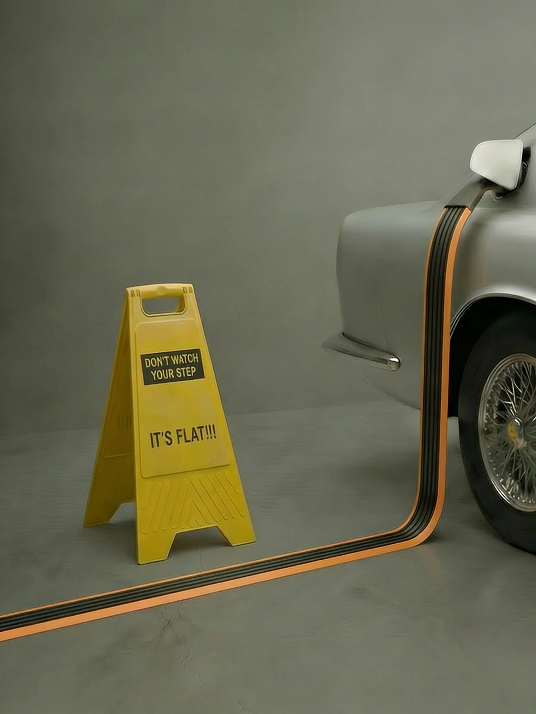 A silver vintage car with a black and orange striped curb along the side, and a yellow caution sign that reads 'Don't watch your step, it's flat!!!' in front of the car.
