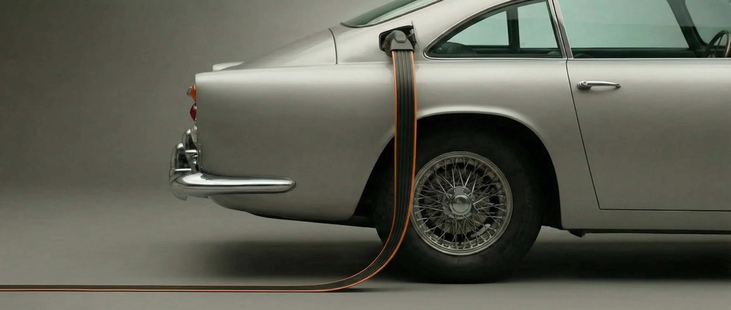 A classic silver car with an electric cord plugged into its rear side, illustrating electric vehicle charging.