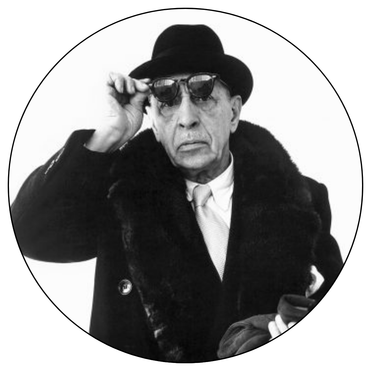 A black and white image of Igor Stravinsky, who is raising his sunglasses to look straight at the camera. He is wearing a black hat and heavy black coat.