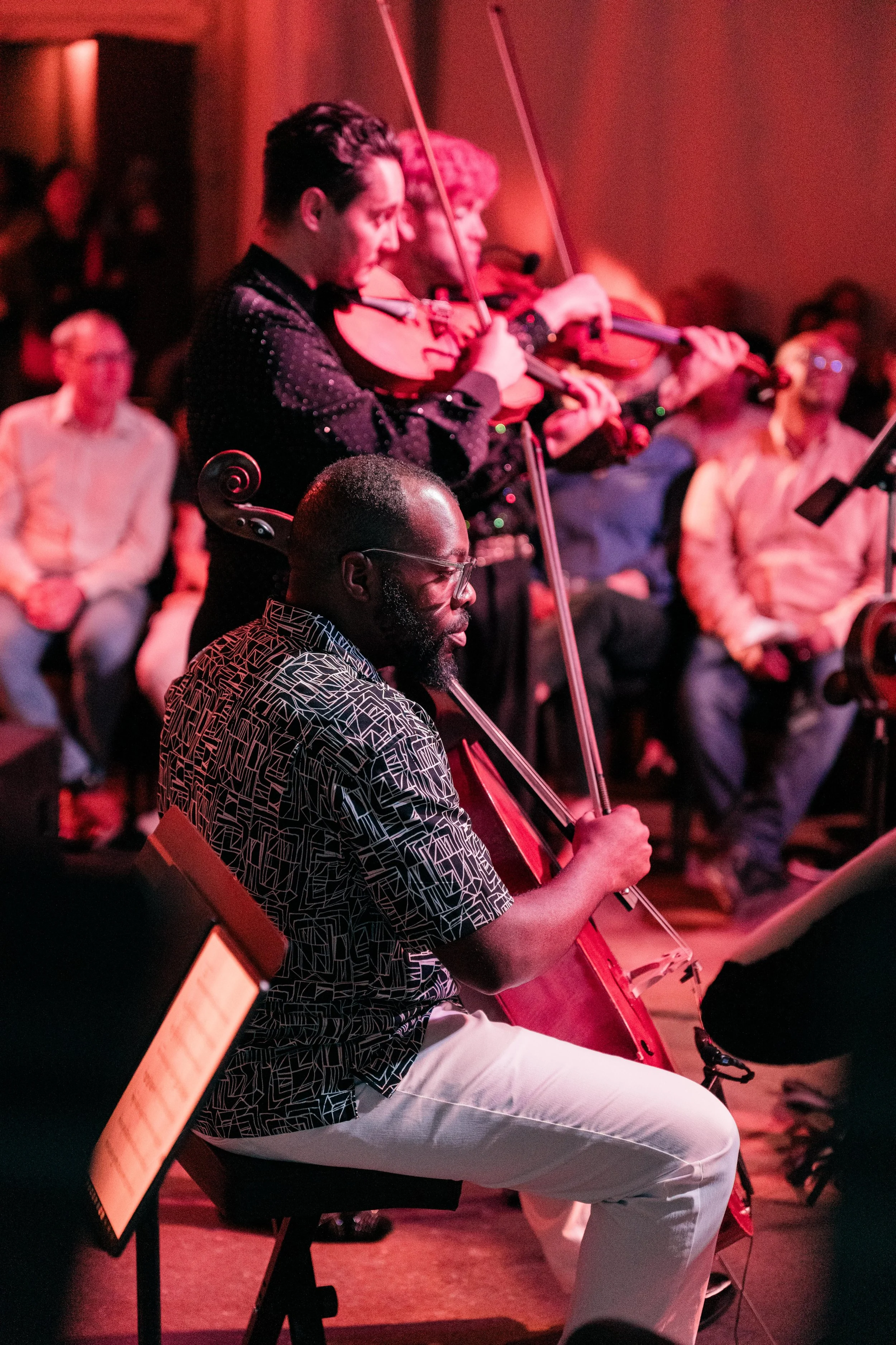 A close-up of a cellist performing with an orchestra, violas and audience in the background