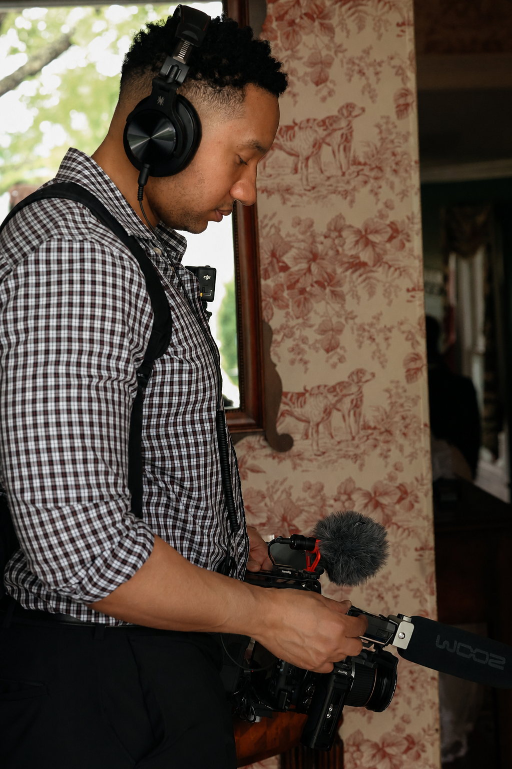 A man with curly black hair wearing a checkered shirt and headphones is holding a professional video camera with a microphone. He is standing indoors near a window with greenery outside, and there is floral wallpaper with dog illustrations on the wall behind him.