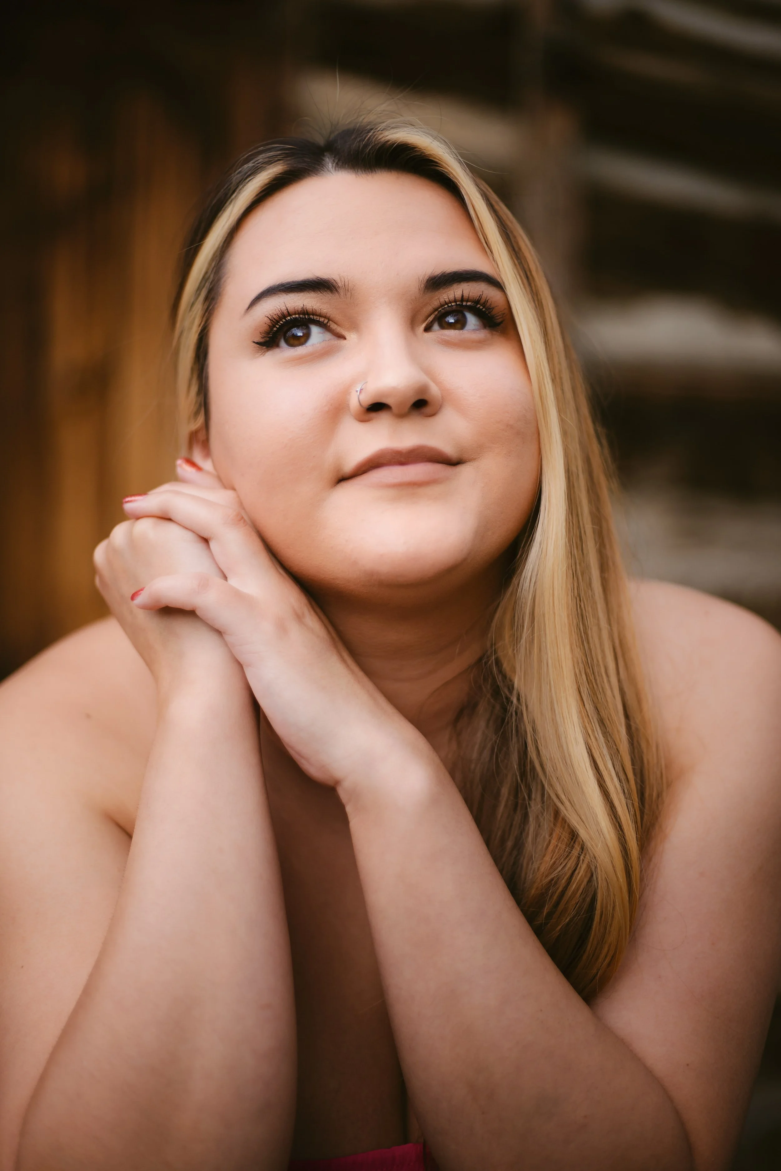 Close-up of a woman with long blonde hair. She is resting her chin on her hands and looking slightly upwards. She has a nose piercing, well-groomed eyebrows, and makeup on her lashes and eyelids. The background is blurred and dark.