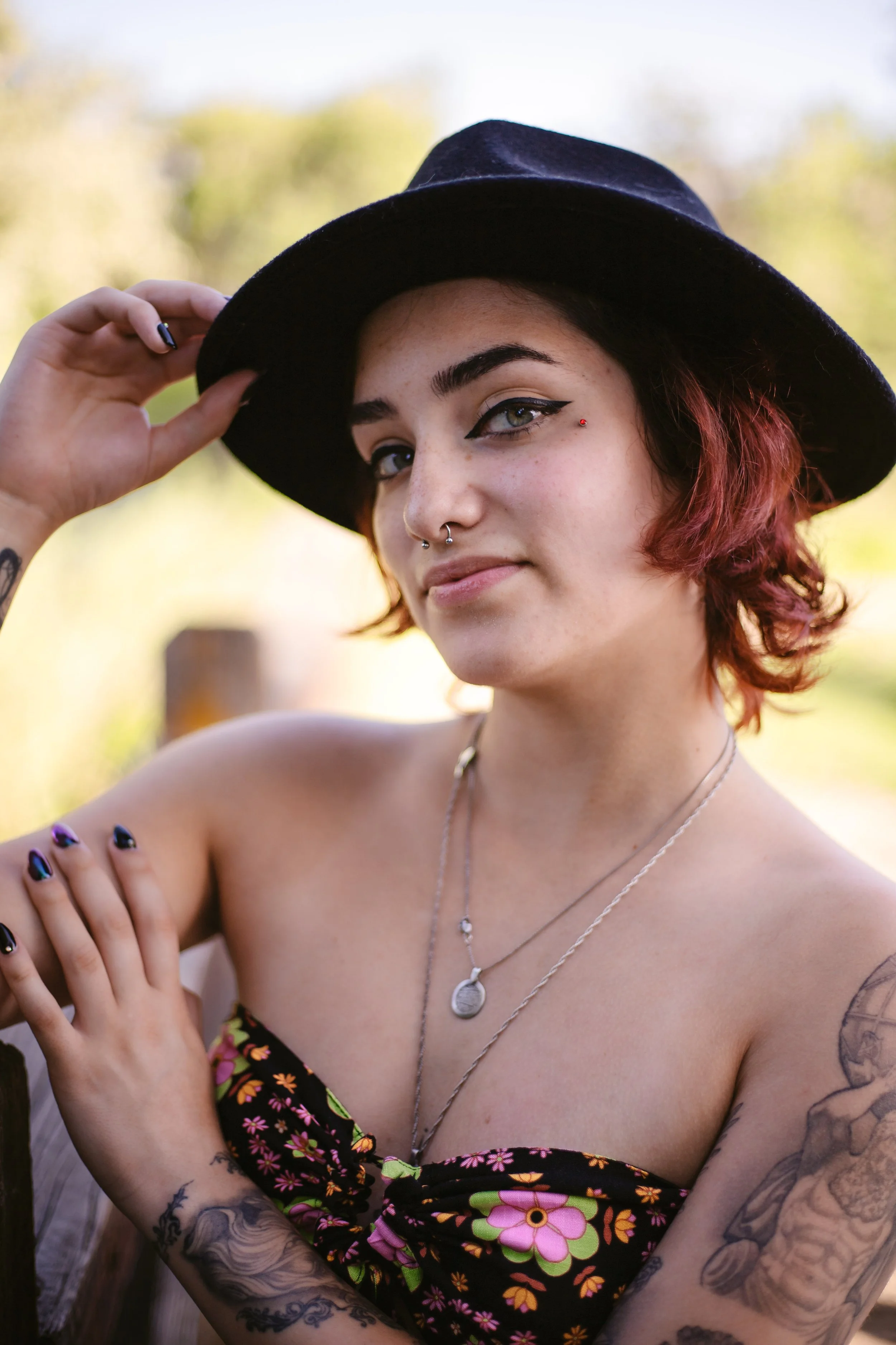 A young woman with short red hair, wearing a black hat, floral dress, and multiple necklaces, is outdoors. She has piercings near her eye and nose, tattoos on her arms, and is holding her hat.