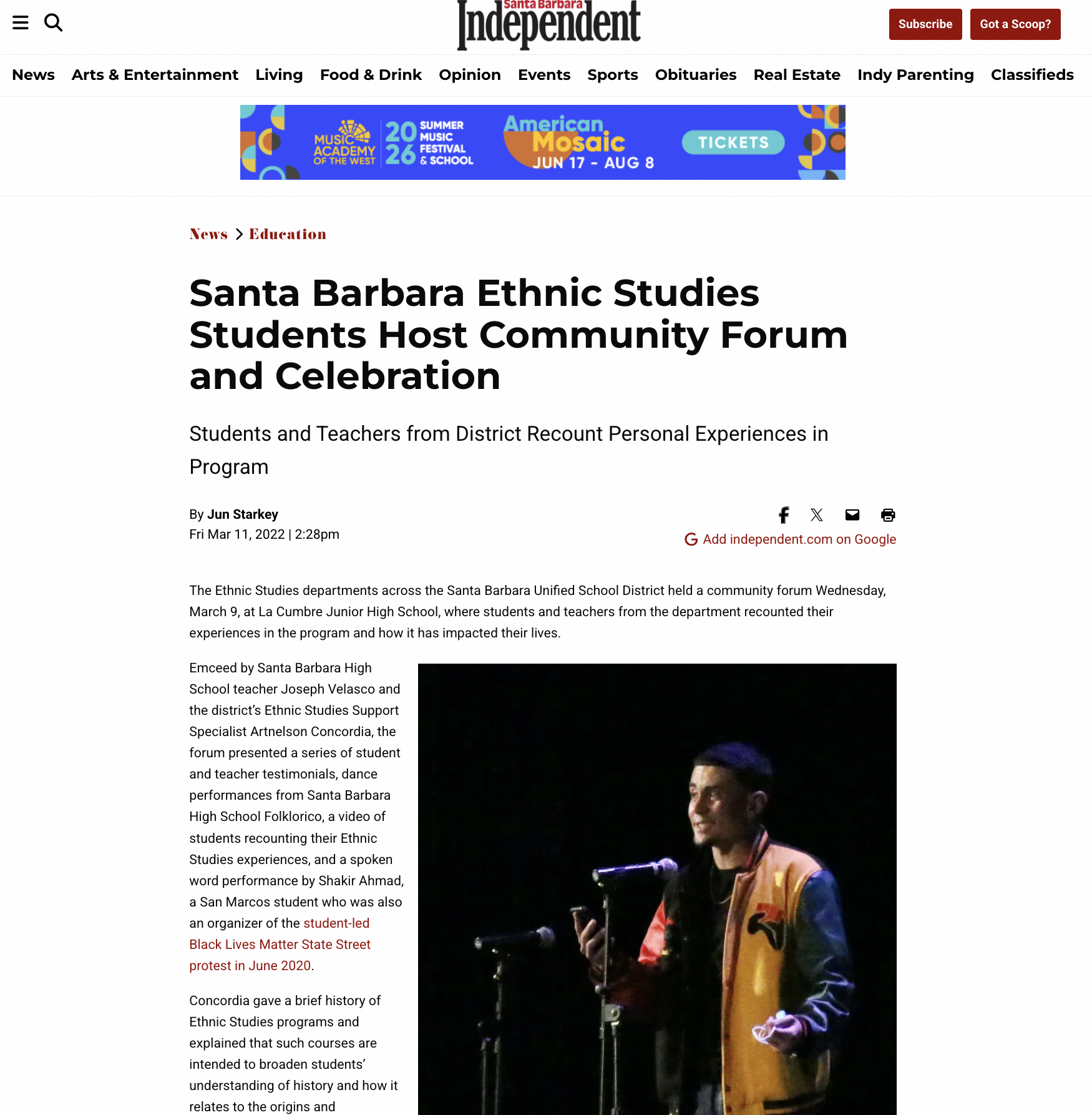 A young man in a colorful jacket standing on a stage during a community forum event, with a microphone and a dark background.