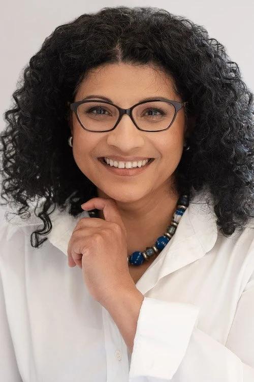 A smiling woman with curly hair wearing glasses, a white blouse, and a beaded necklace.