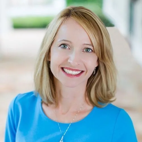 Portrait of a woman with shoulder-length blonde hair, wearing a blue top, smiling and looking at the camera with a blurred outdoor background.