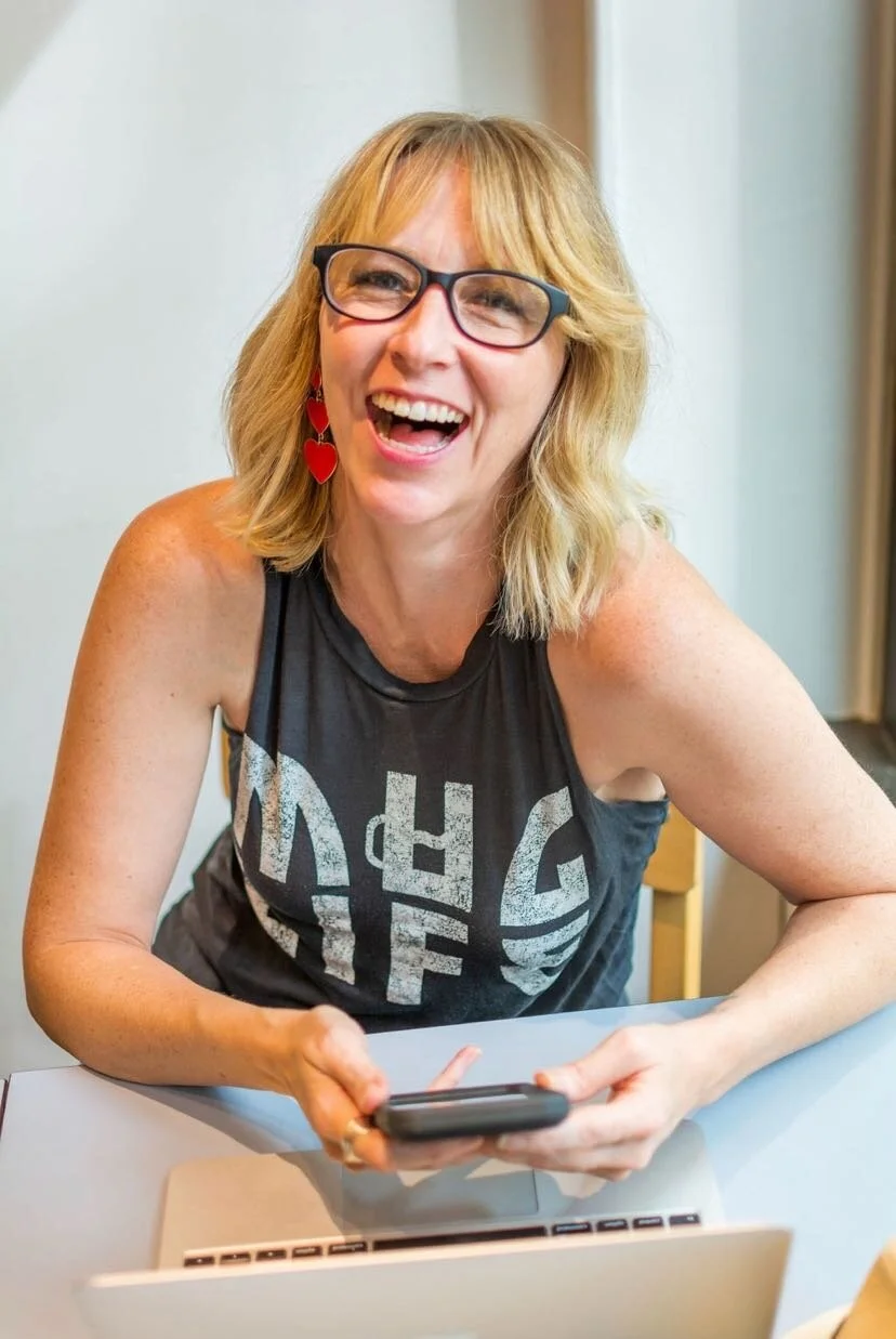 Smiling woman with blonde hair and glasses, wearing heart earrings, holding a smartphone, sitting at a table with a laptop.