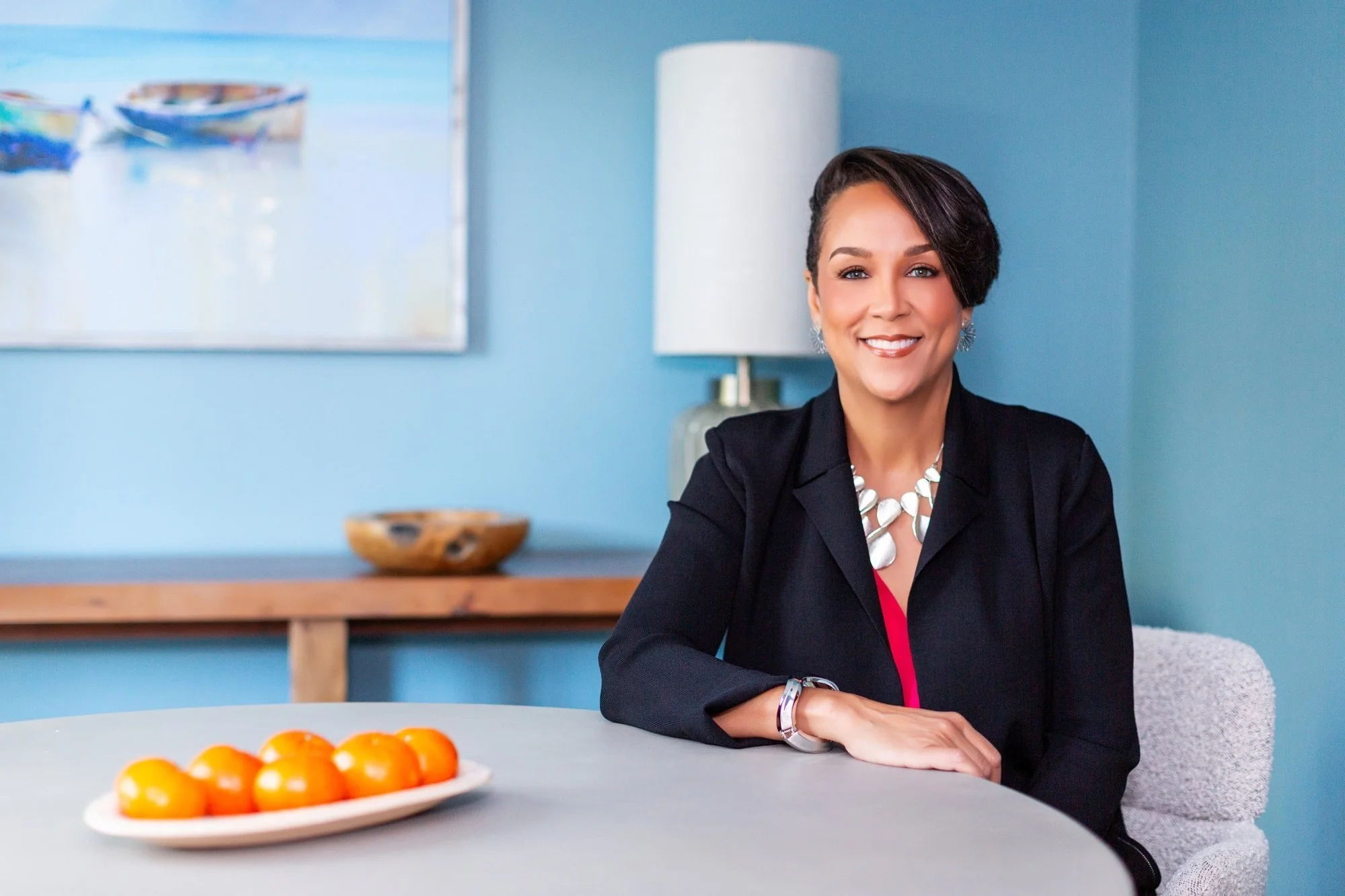 Woman with short dark hair wearing a black blazer and white necklace, sitting at a table with a plate of oranges, in a bright room with blue walls and a painting of boats.