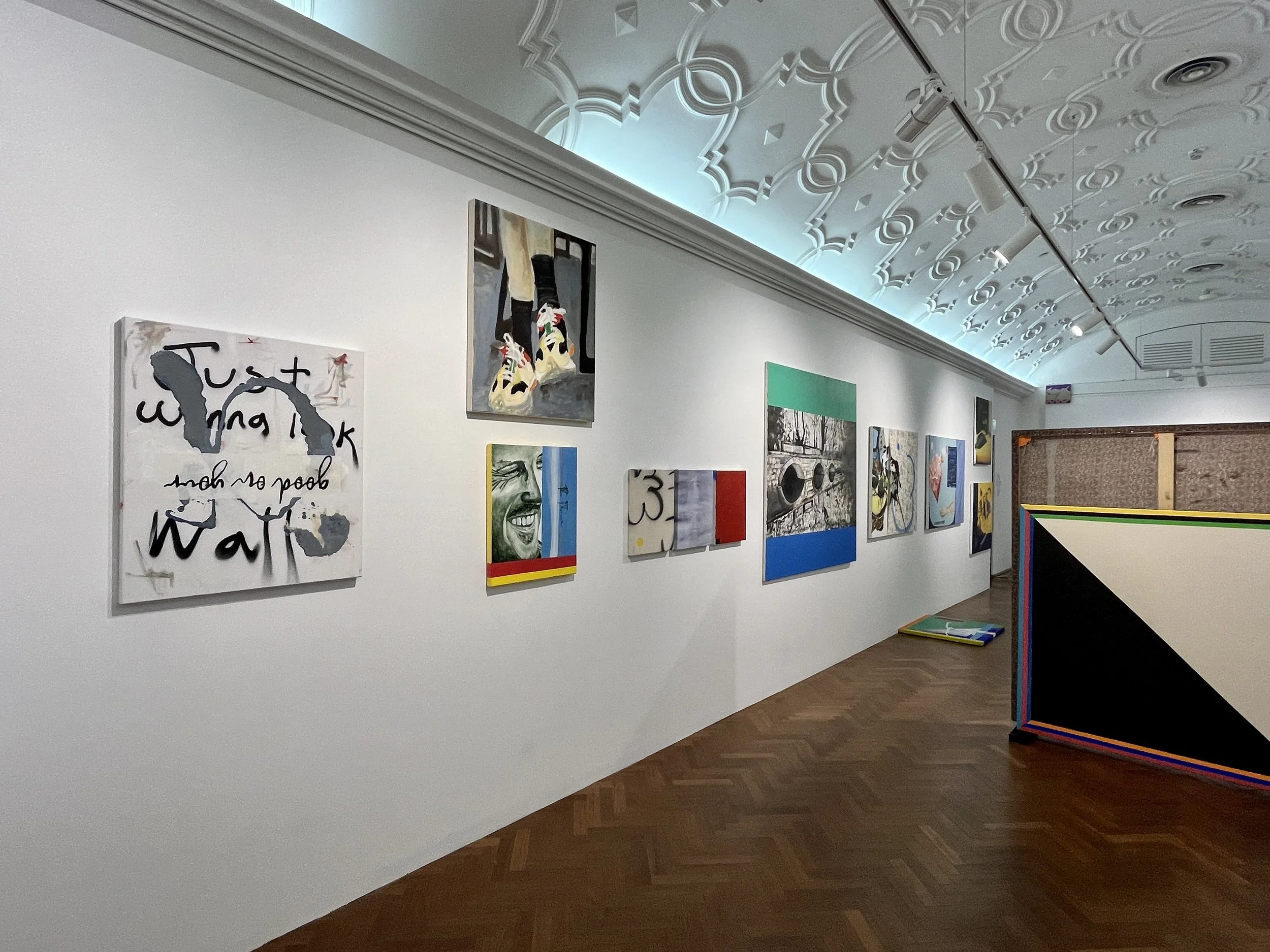 An art gallery with multiple colorful paintings on a white wall, featuring various styles and subjects, with a white ceiling that has intricate decorative molding and track lighting.
