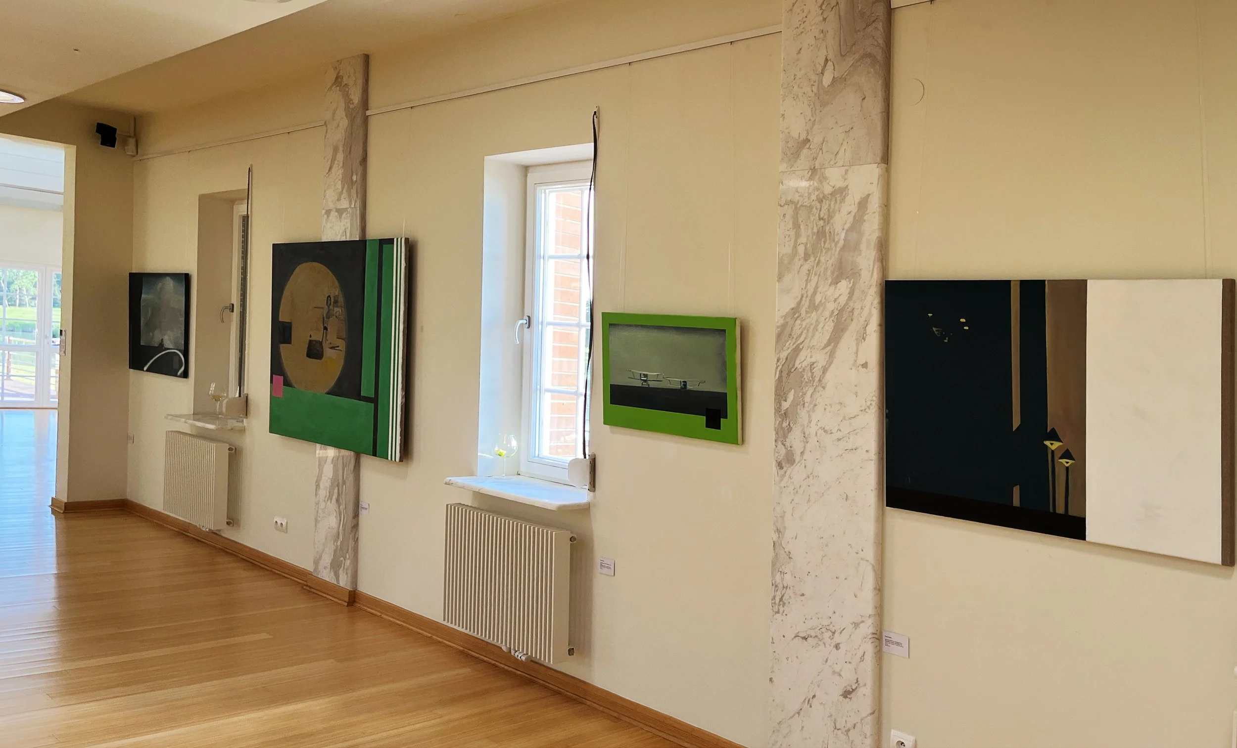 An art gallery wall with several paintings, including one with a green frame, in a room with wooden floors and beige walls with marble accents.