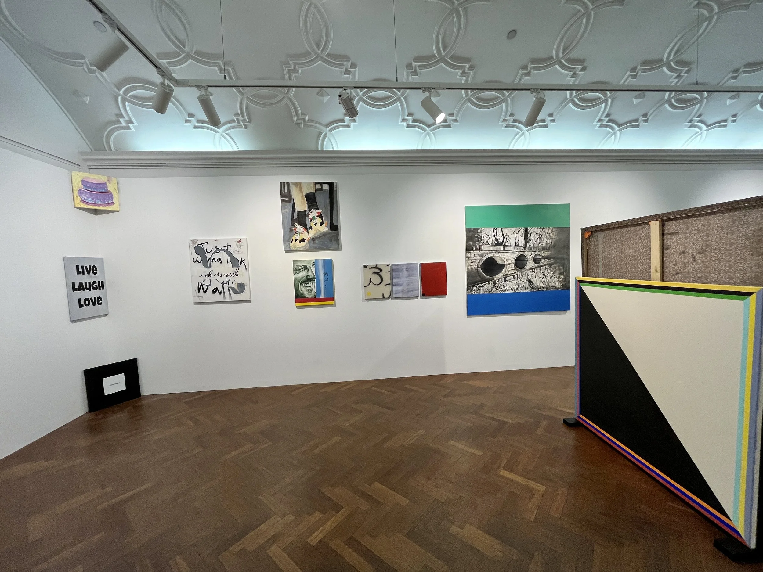 An art gallery with white walls displaying various paintings including a colorful shoe, a smiling face, and abstract art. A black frame is on the floor. The ceiling has a decorative pattern with spotlights.