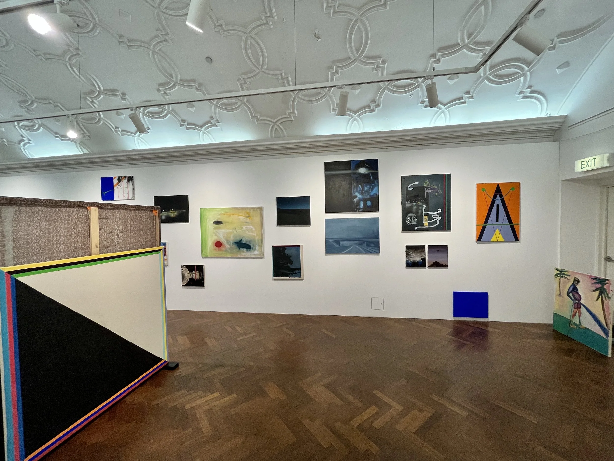 An art gallery with various paintings hung on white walls. The ceiling has intricate molding and track lighting, and the wooden floor adds warmth to the space.