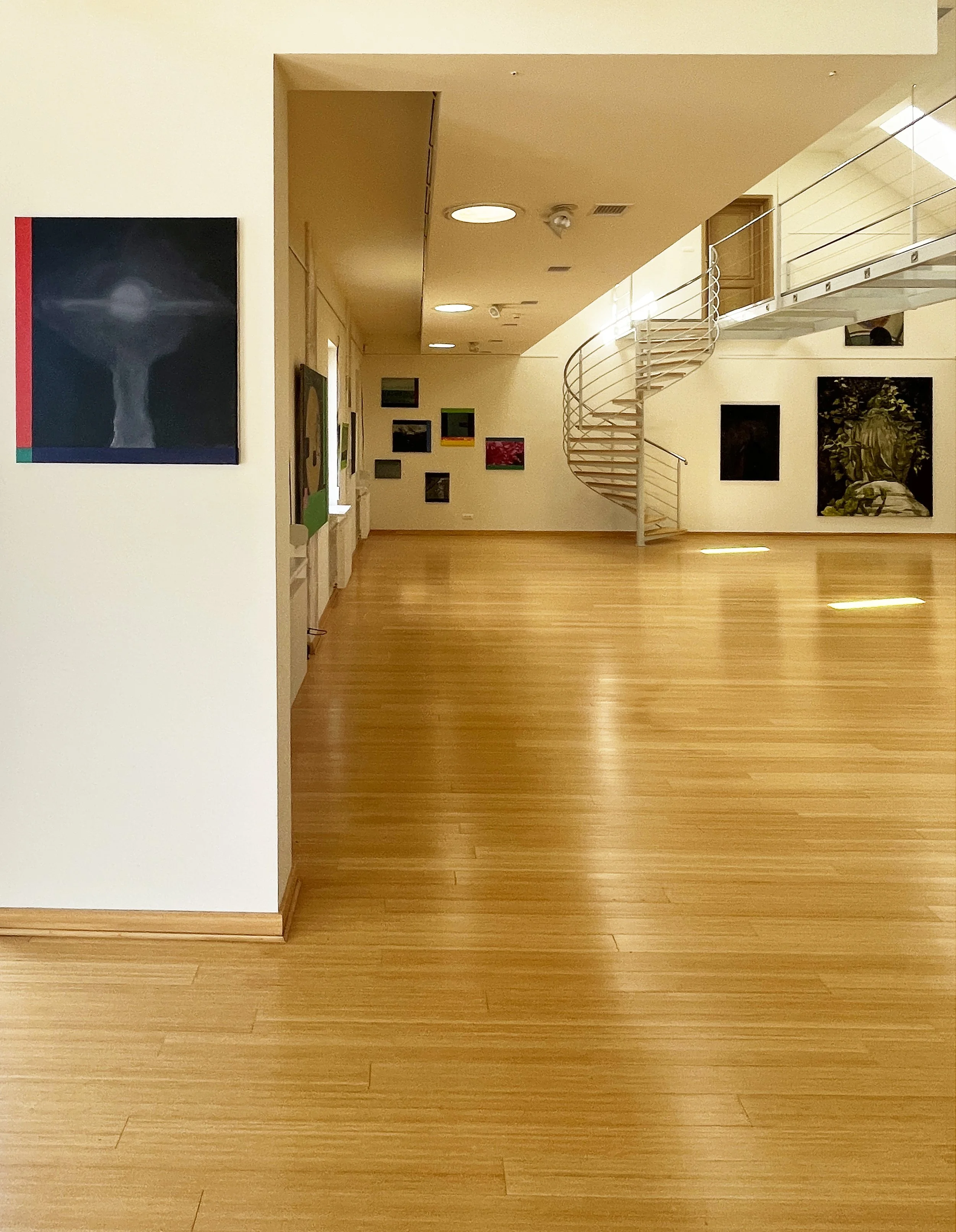 An art gallery interior with framed paintings on cream-colored walls, a modern spiral staircase, wooden flooring, and ceiling lights.