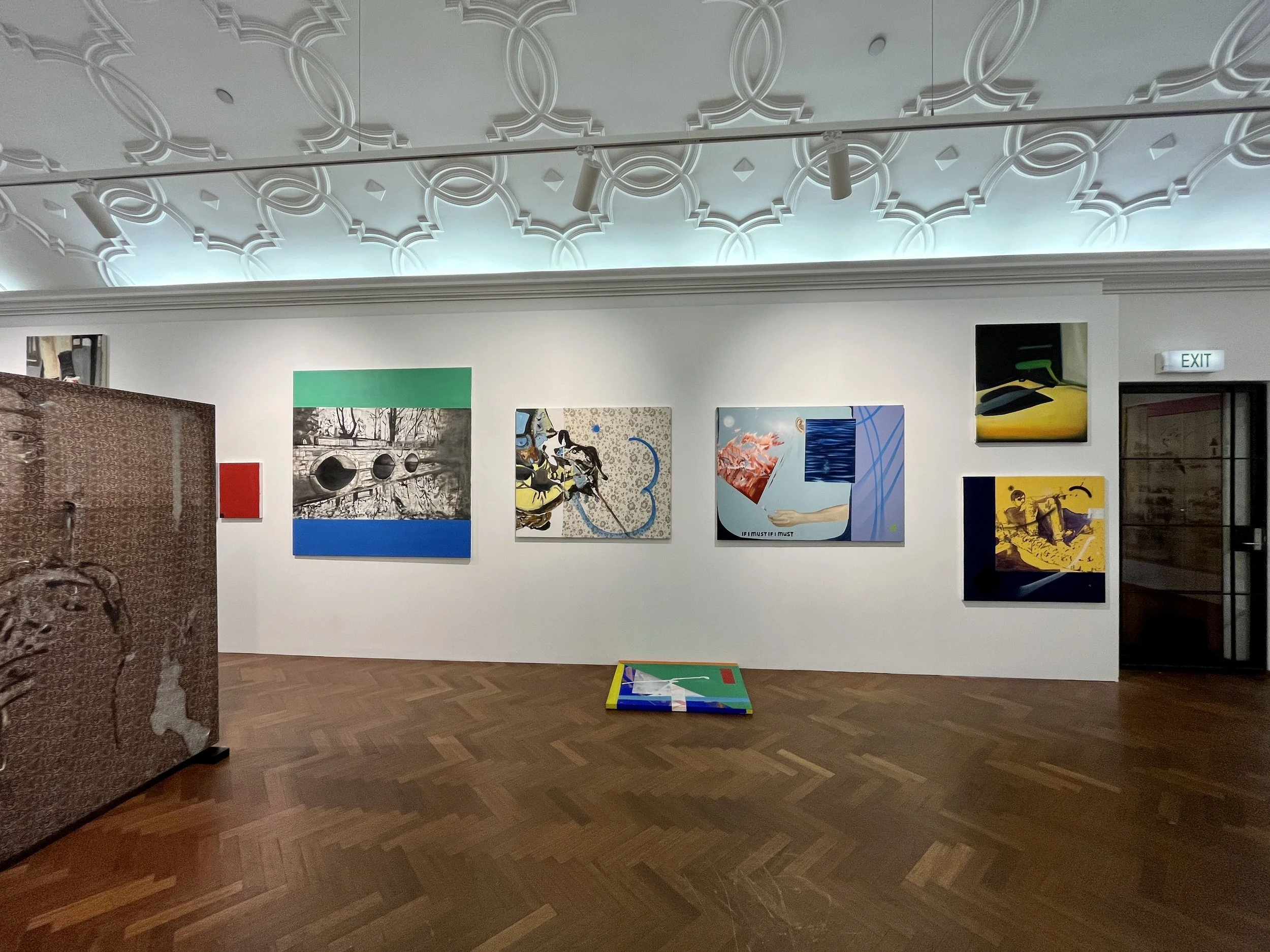 An art gallery with various colorful paintings on white walls and a patterned ceiling with track lighting, a wooden floor, a patterned fabric panel on the left, and a small table or chessboard on the floor.