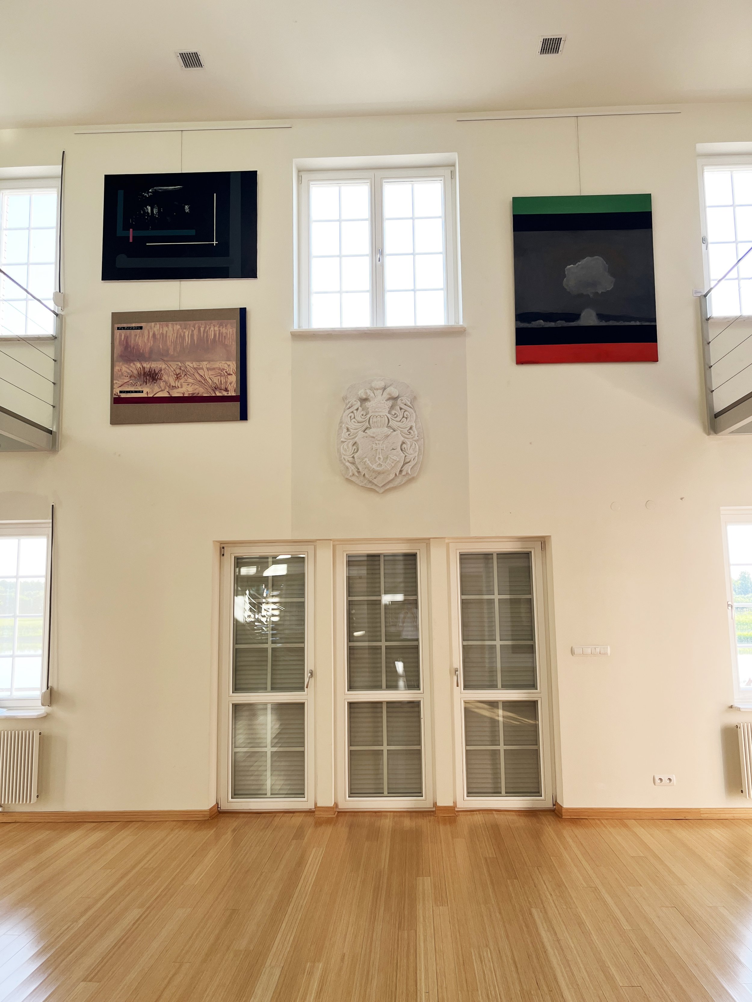 An interior room with three large windows, a white wall with artwork, a decorative shield, and wooden flooring.