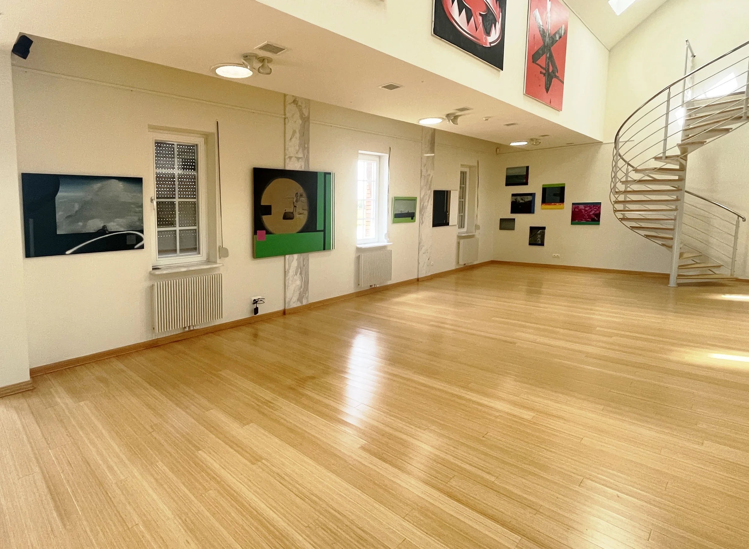 An art gallery interior with various colorful paintings on the wall, hardwood flooring, and a spiral staircase to the right.