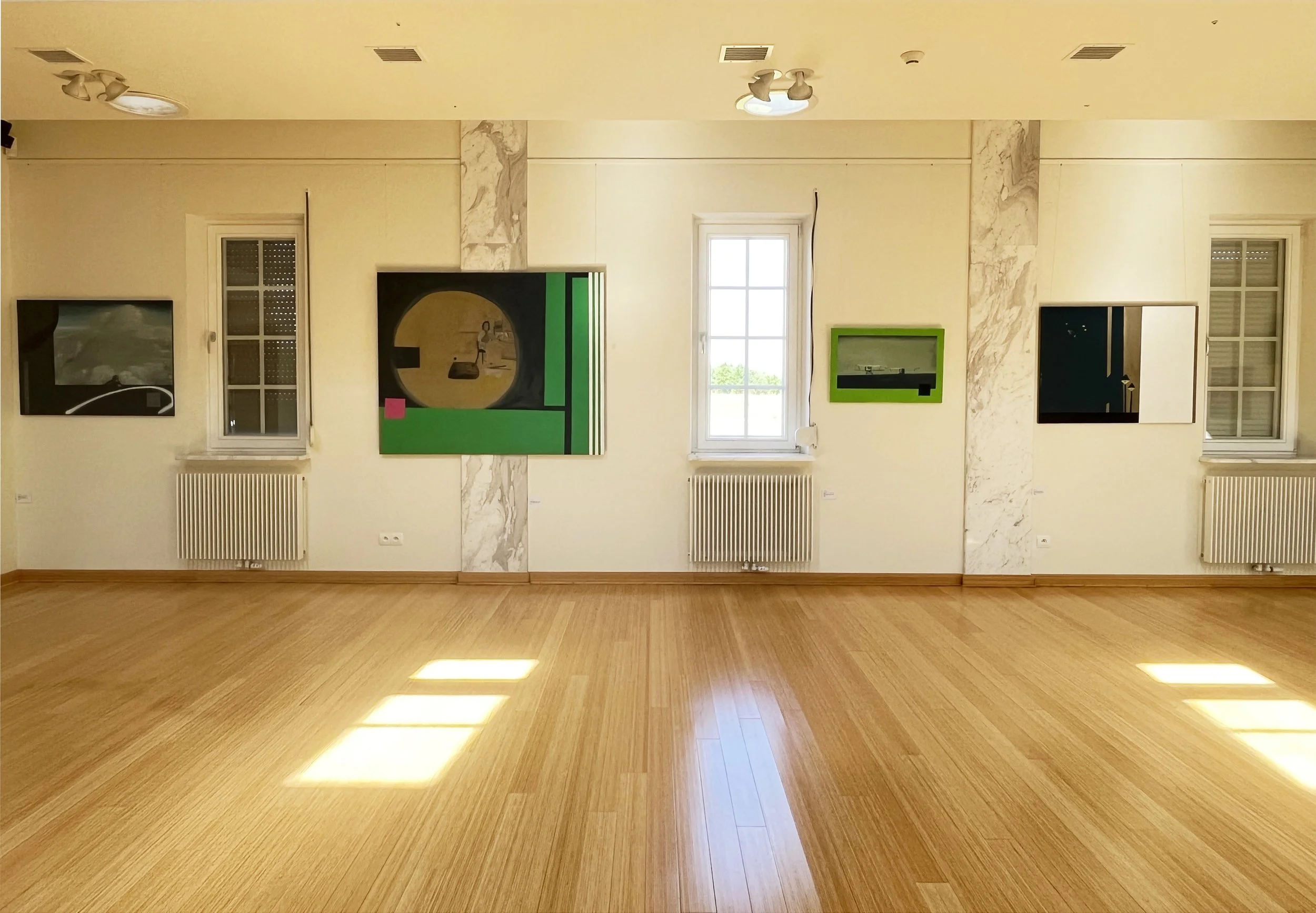 Art gallery with four framed abstract artworks on a light-colored wall and two windows, wooden floor, ceiling lights, and radiators beneath the windows.
