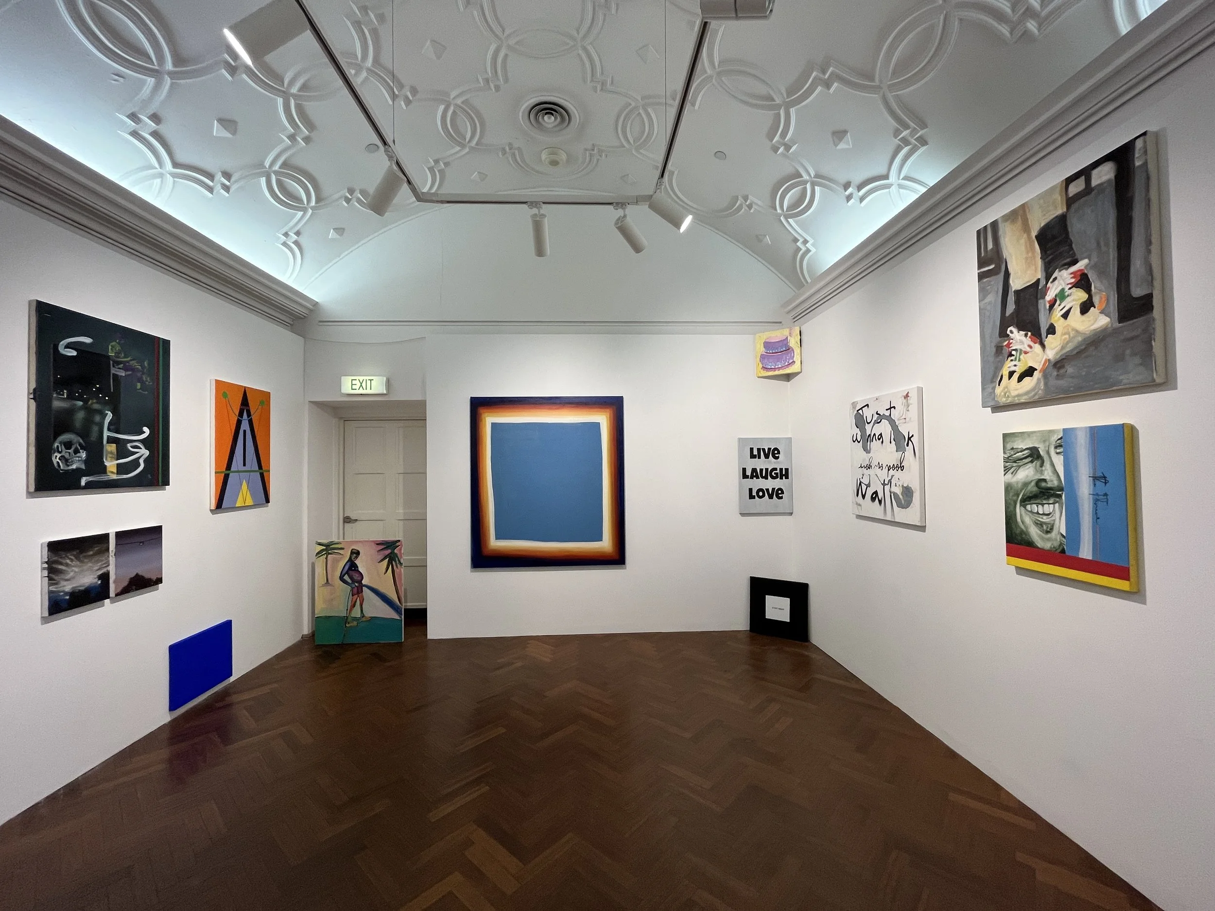 An art gallery with white walls displaying various colorful paintings, including portraits, abstract art, and text-based artwork. The ceiling has intricate molding and track lighting, and the floor is wooden with a herringbone pattern.