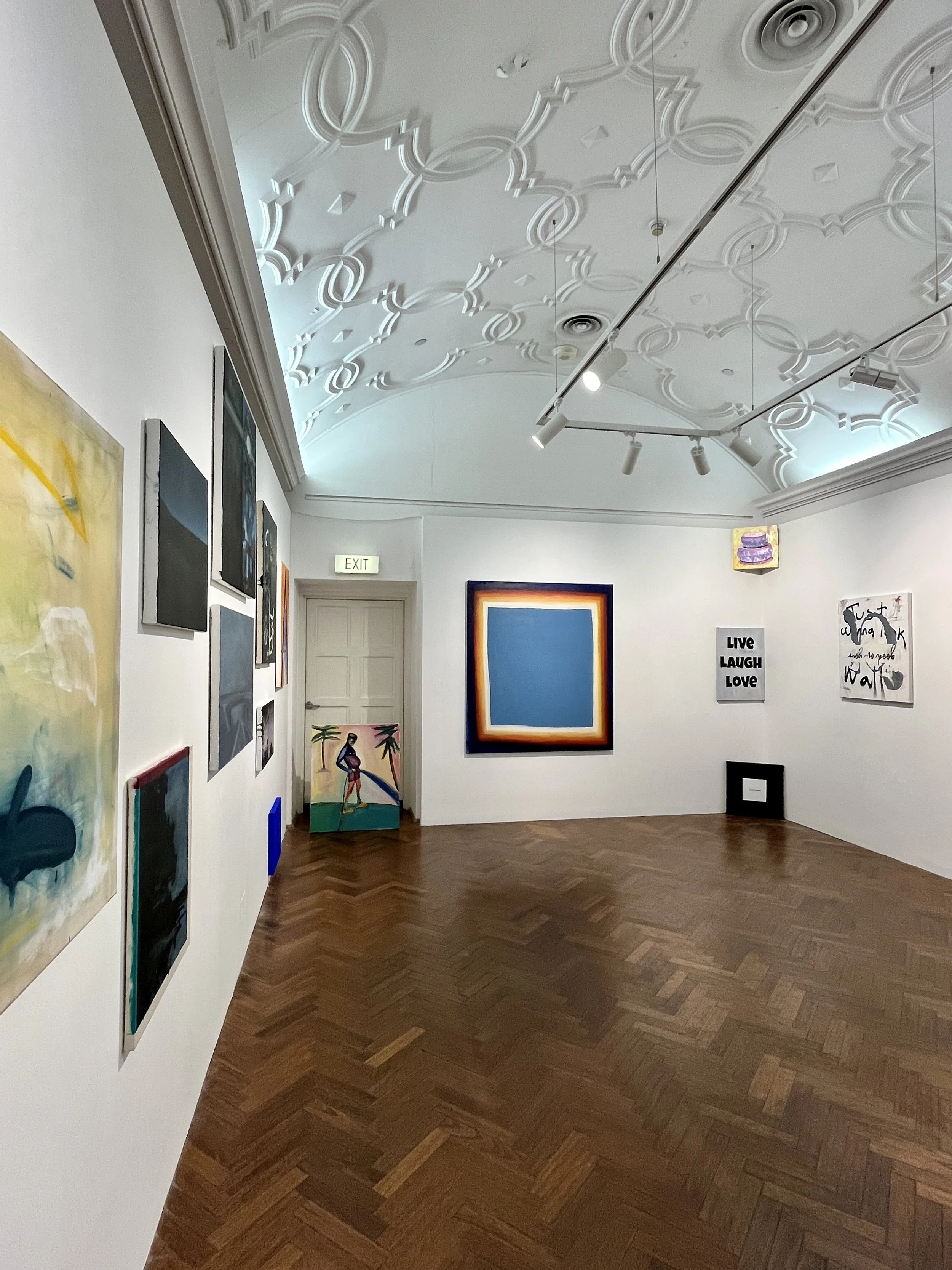 An art gallery gallery featuring various contemporary paintings displayed on white walls. The ceiling has intricate decorative molding and track lighting highlighting the artwork. The floor is wooden in a herringbone pattern, and there is a green exi